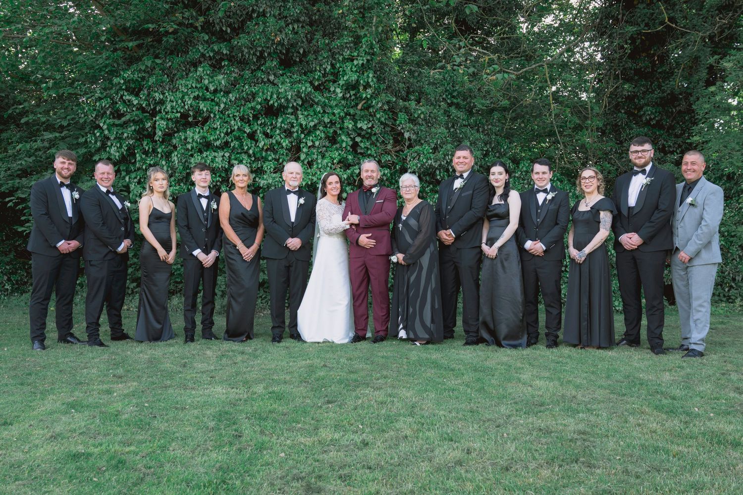 Wedding party in formal attire posing outdoors on green lawn with lush tree backdrop.