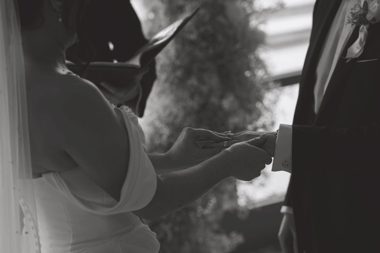 Black and white wedding photo of bride and groom exchanging rings during outdoor ceremony.