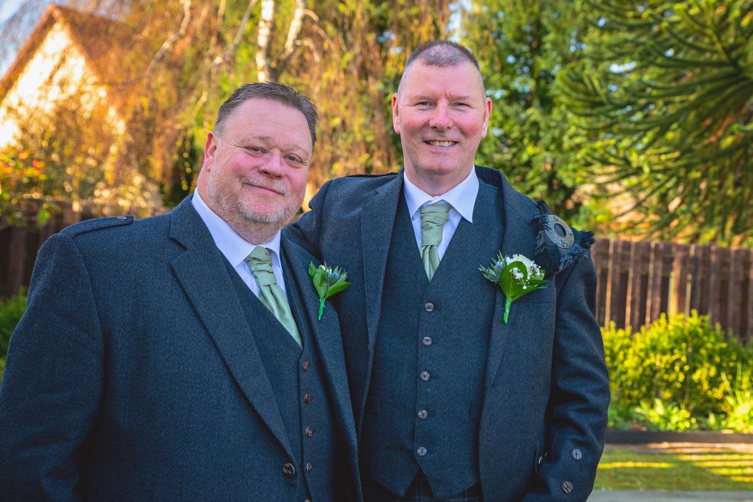 Groomsmen in traditional Scottish kilts and suits gather for formal photos in a sunny garden.