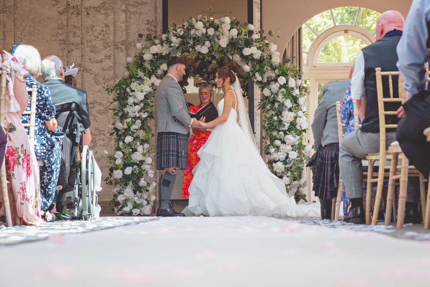 Wedding ceremony under a floral arch with guests seated on either side.