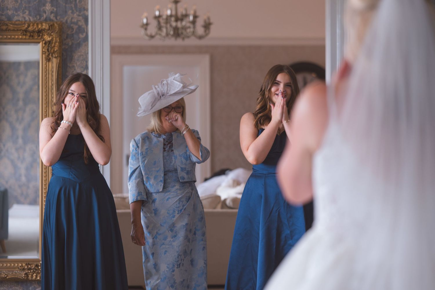 Bridesmaids in blue dresses and mother of the bride react emotionally seeing bride in wedding dress reflected in mirror.