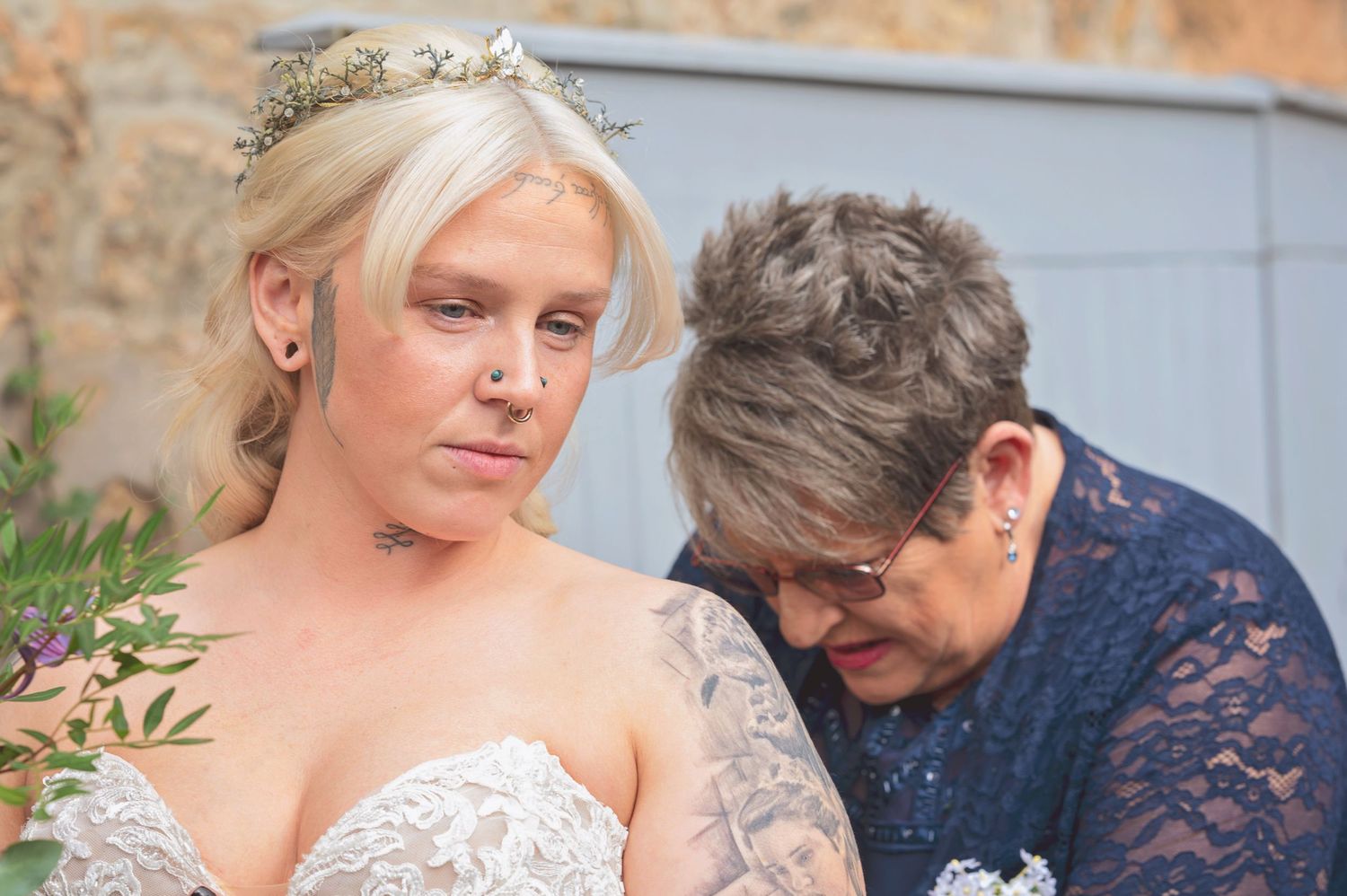 Emotional bride in lace gown with floral crown receives comfort from older woman at wedding.