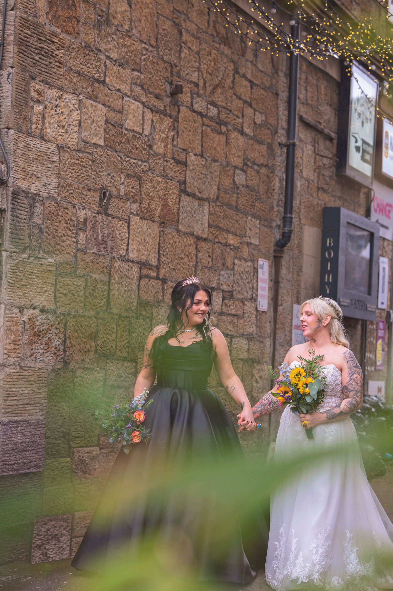 Two brides hold hands outside a stone wall, one in a black gown and one in a white lace dress.