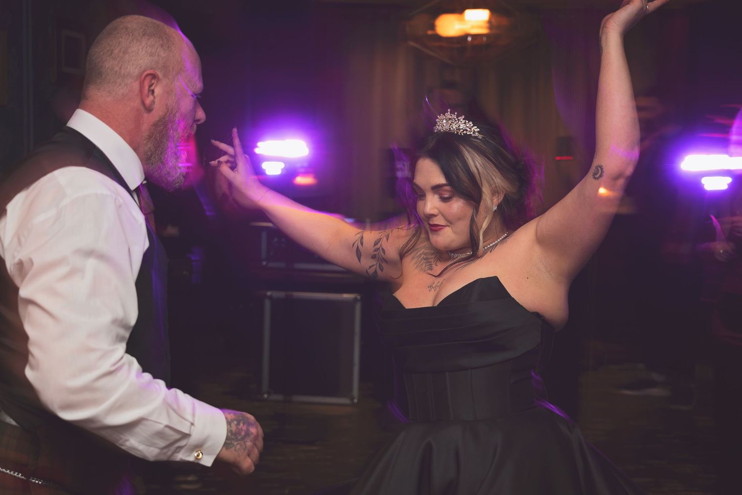 A woman in a black ballgown and tiara dances energetically with a man at a wedding reception.