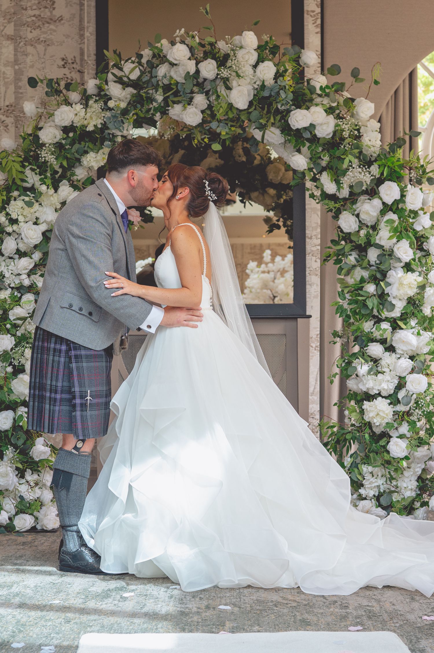 Wedding couple shares intimate moment under white rose floral archway.