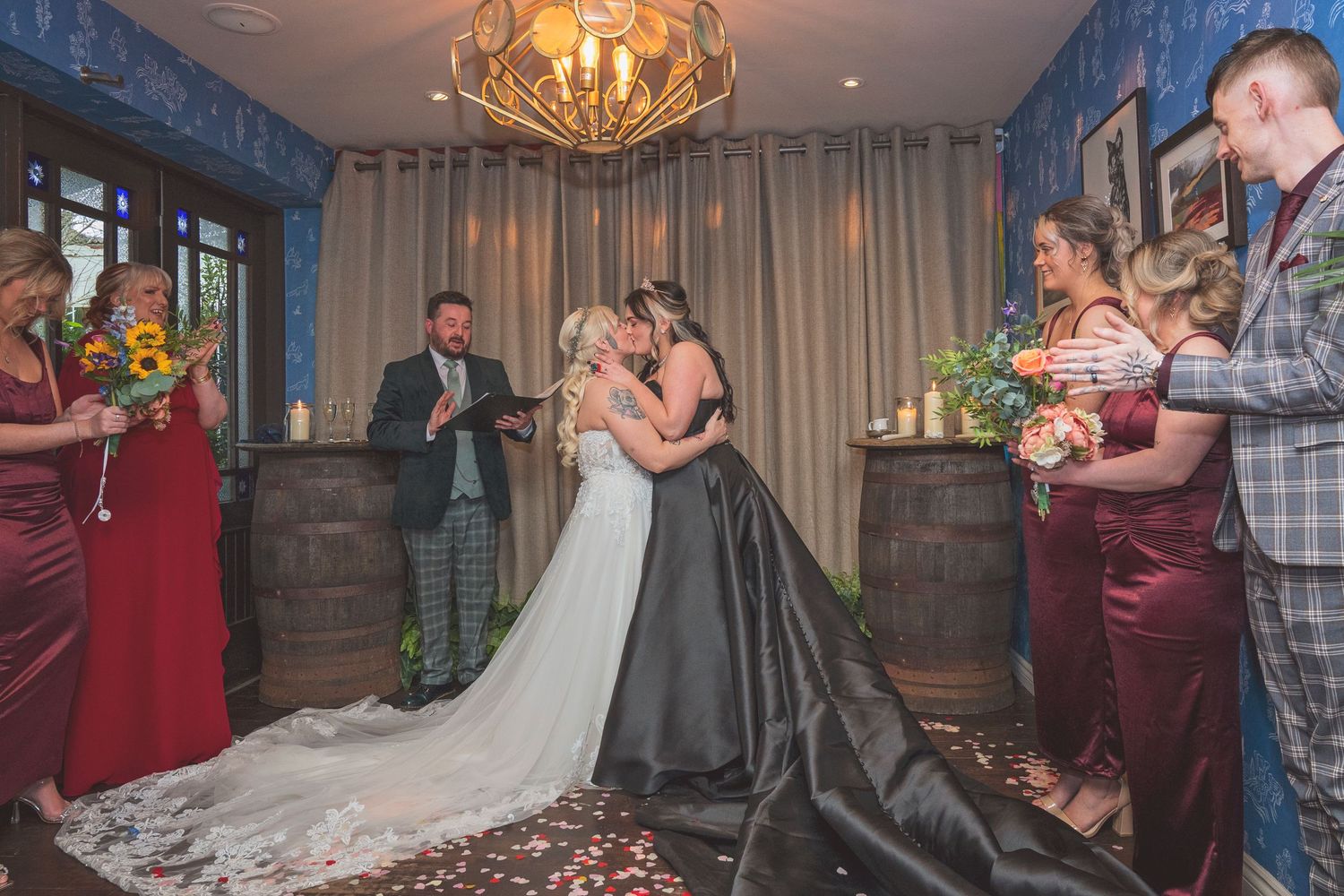 Two brides share their first kiss surrounded by wedding party members in a warmly lit ceremony venue.