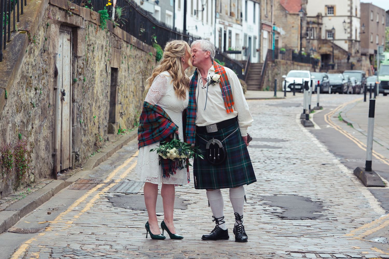 Couple in Scottish attire sharing a kiss on cobblestone street, groom in tartan kilt, bride in white dress with plaid shawl.