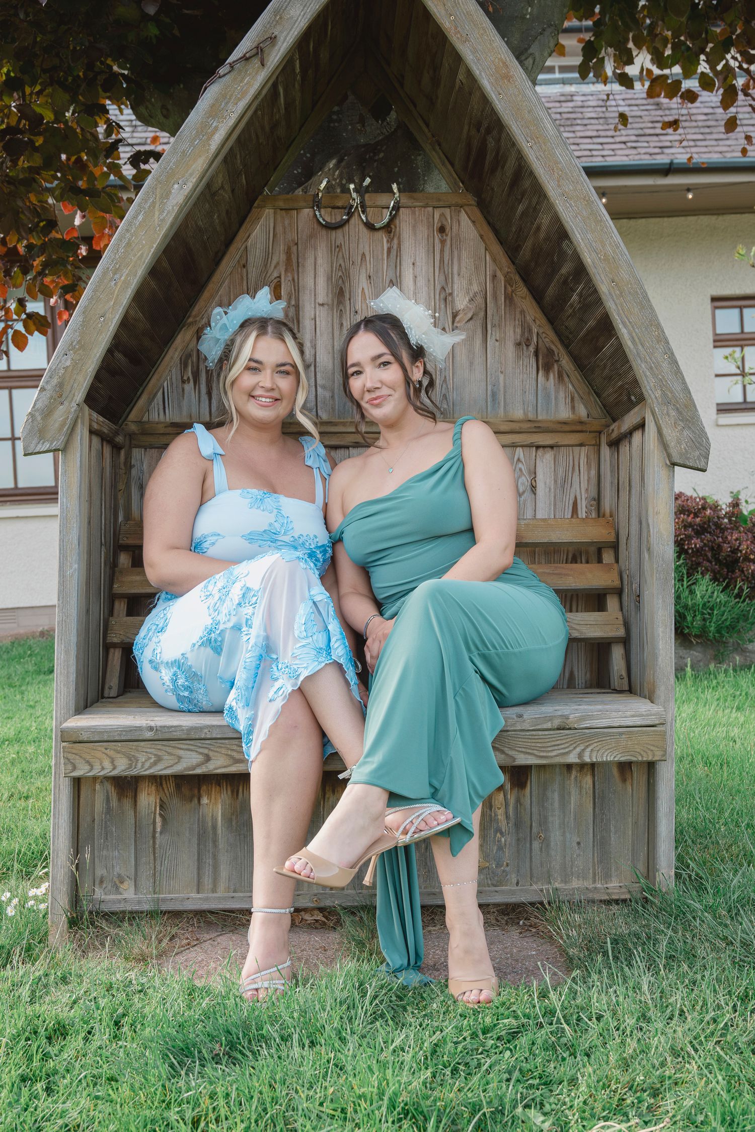 Two women in elegant dresses sitting in a wooden garden arbor bench, smiling outdoors.