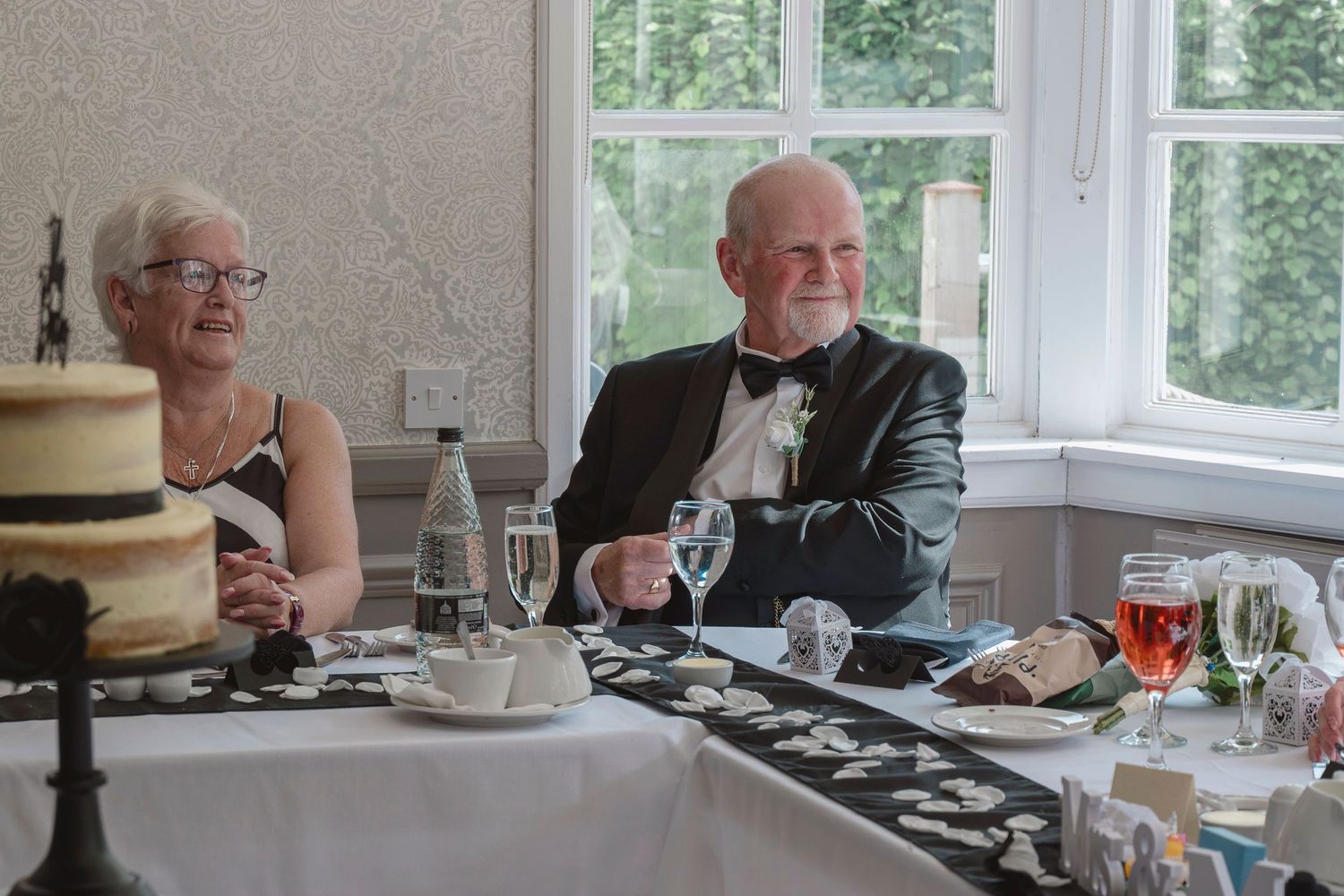 Elderly couple in formal attire seated at a wedding reception table with cake, glasses, and elegant decor.