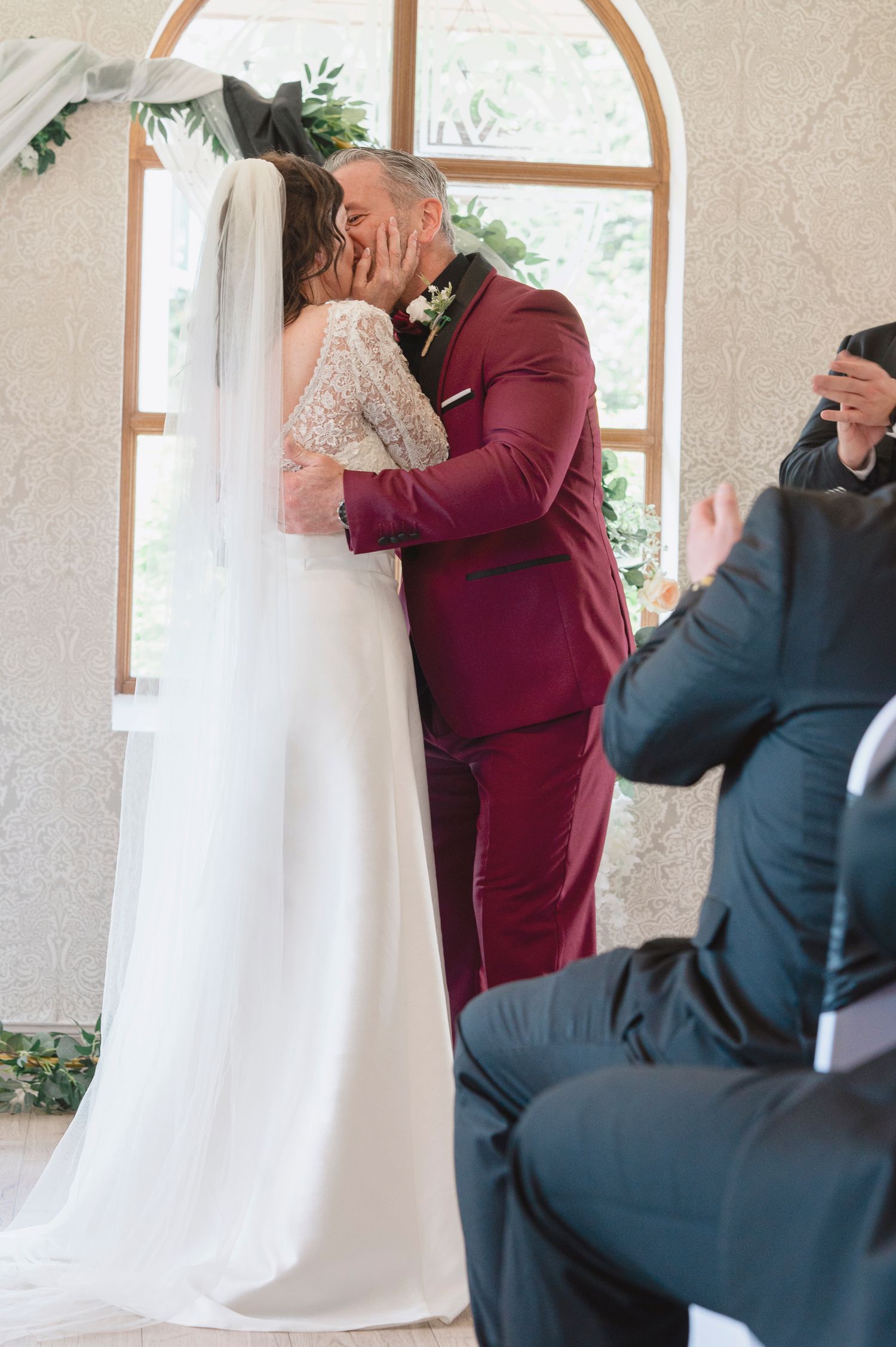 Bride and groom share their first kiss at the altar during a wedding ceremony, groom wearing a burgundy suit.