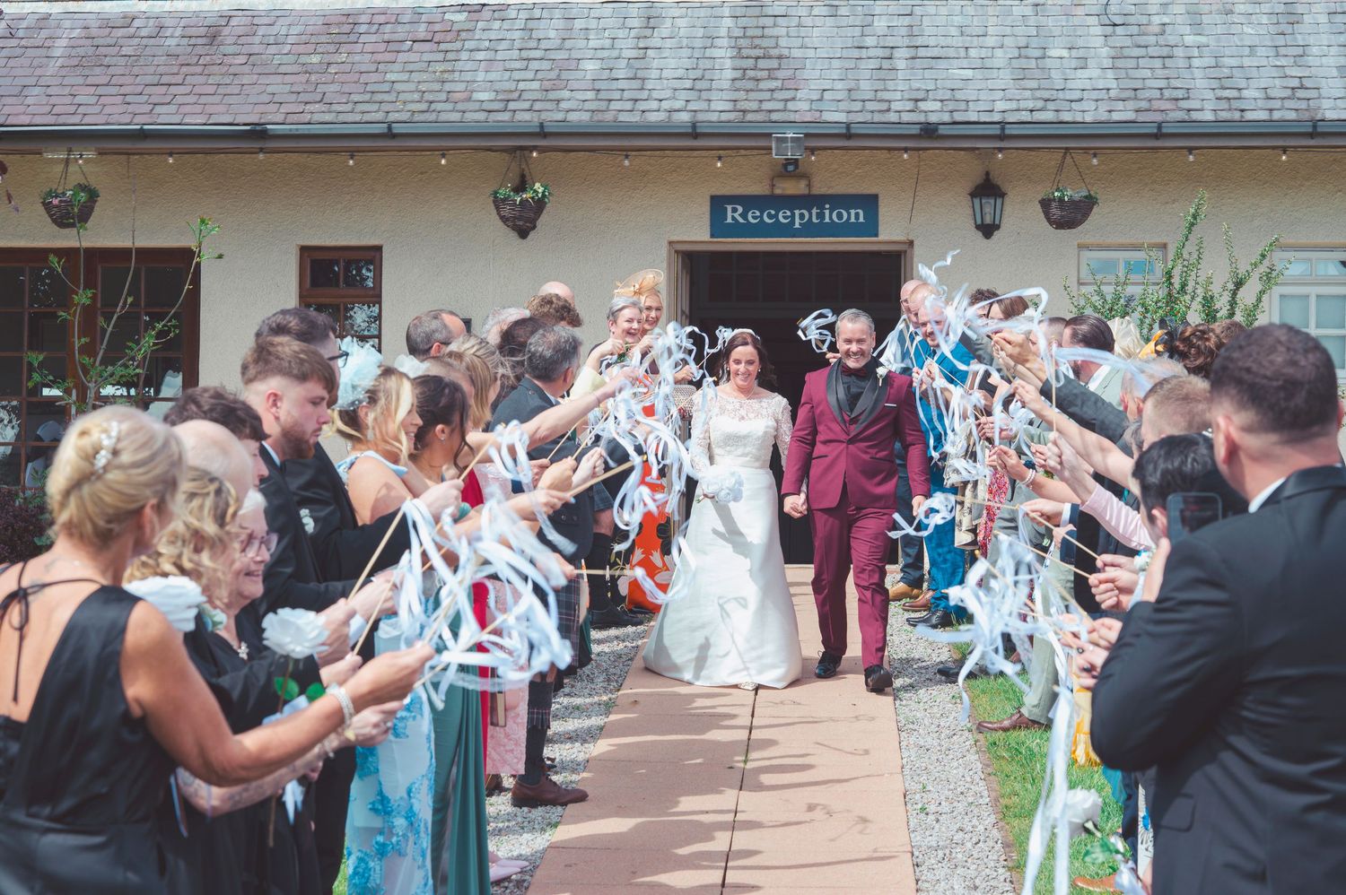 Bride and groom exit wedding reception venue through a ribbon wand send-off with smiling guests lining the path.