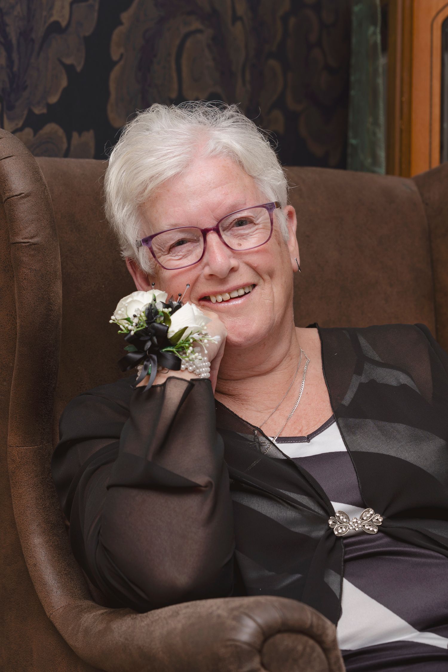 Smiling elderly woman with white hair and purple glasses seated in a leather chair holding white roses.