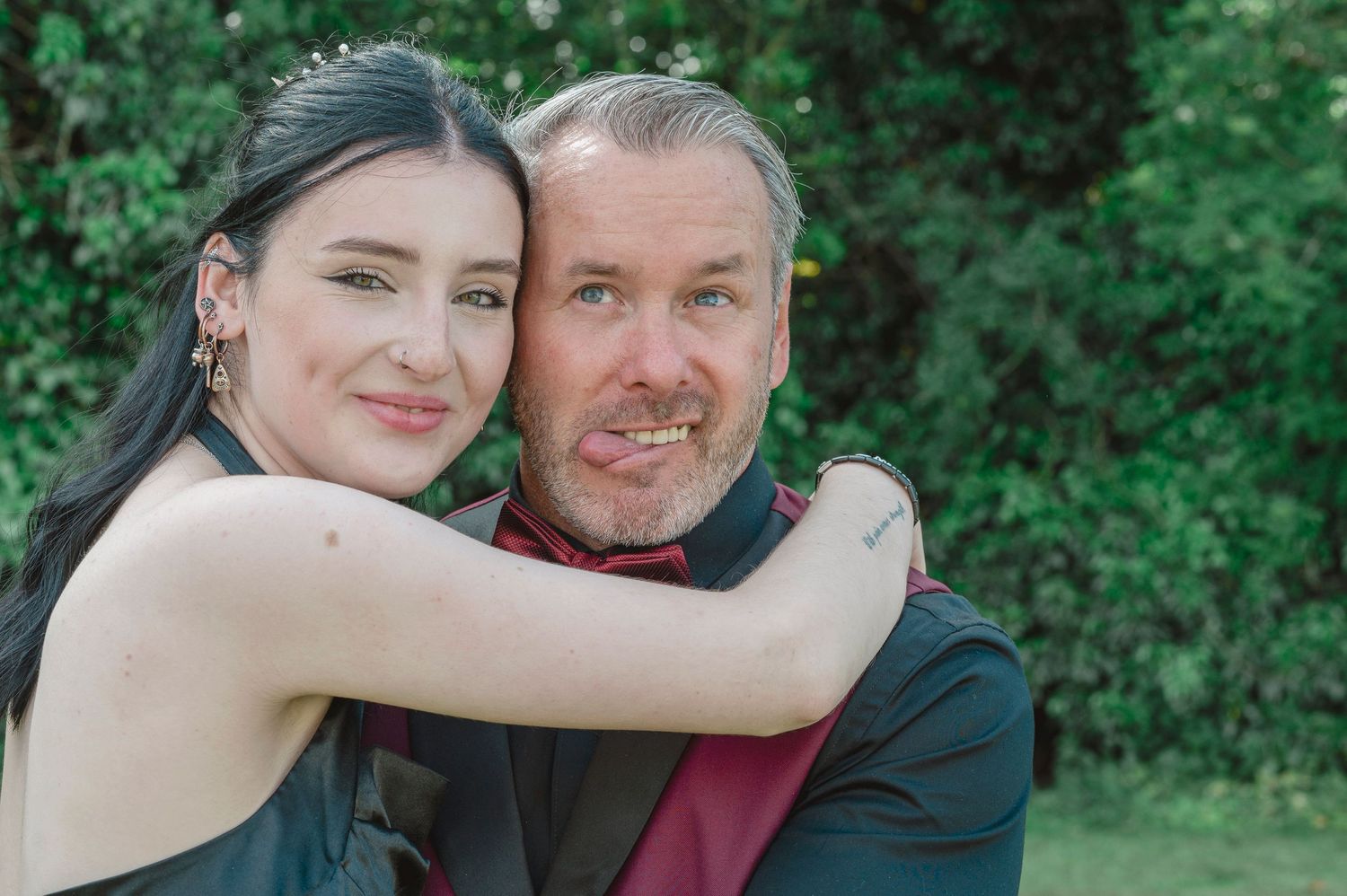 A smiling couple poses outdoors, woman in black dress hugging man in dark suit with red shirt, green trees behind.