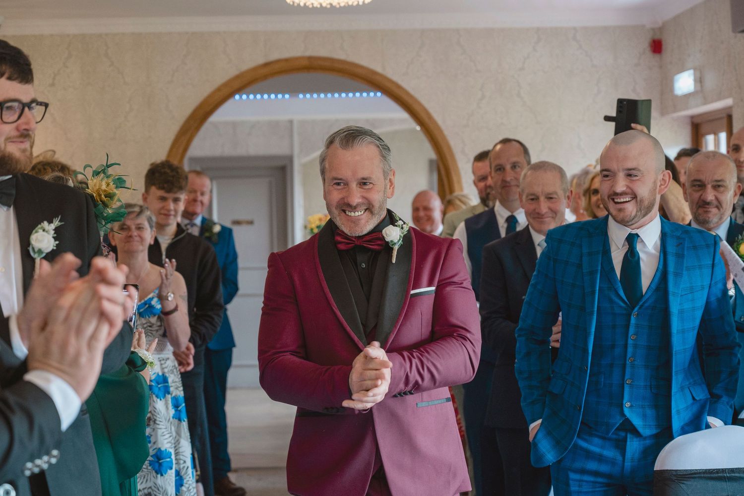 Groom in burgundy tuxedo smiling at wedding ceremony surrounded by guests in formal attire.