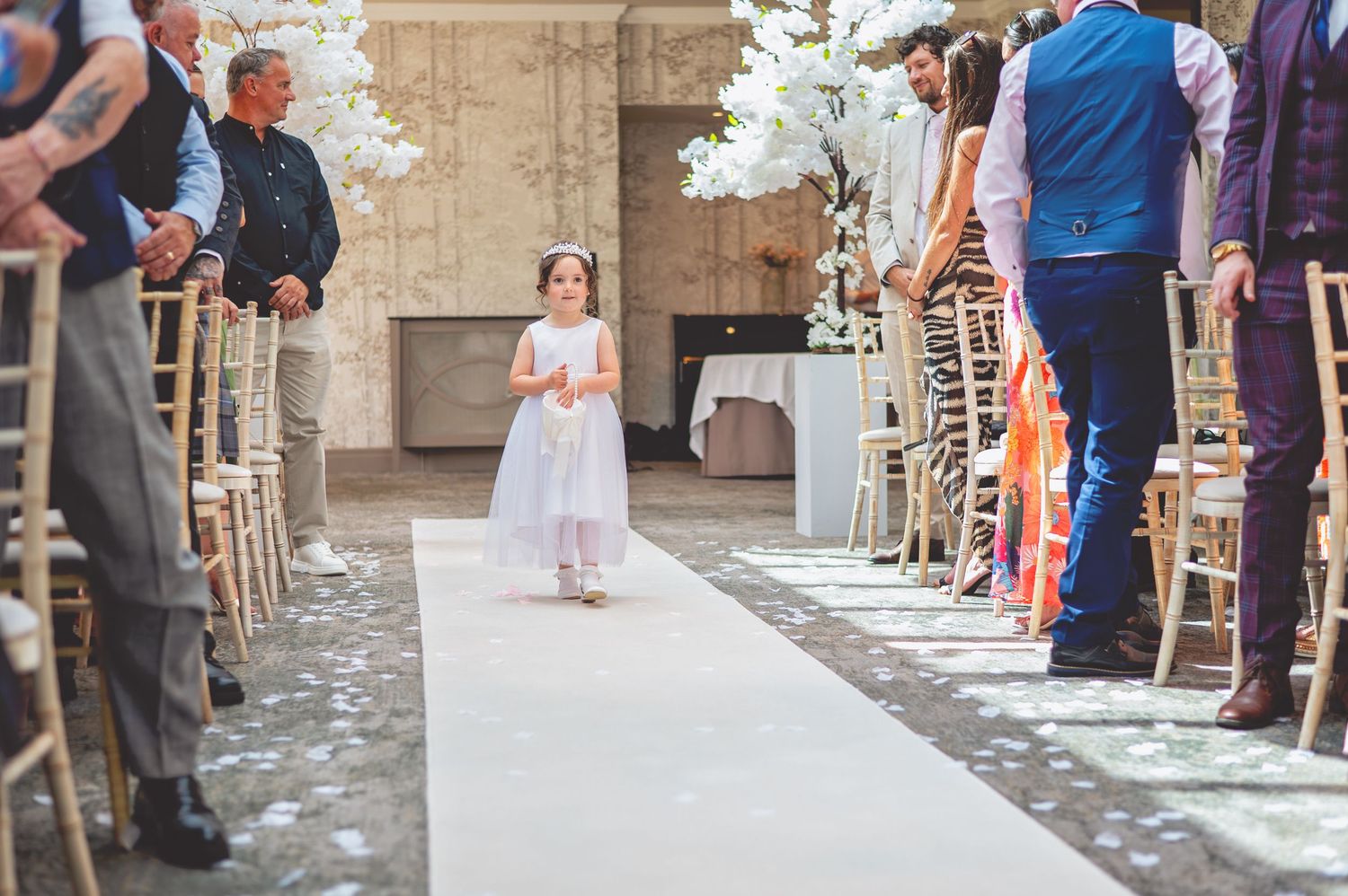 A young attendant in a white dress walks down a white aisle runner lined with decorated chairs at a wedding ceremony.