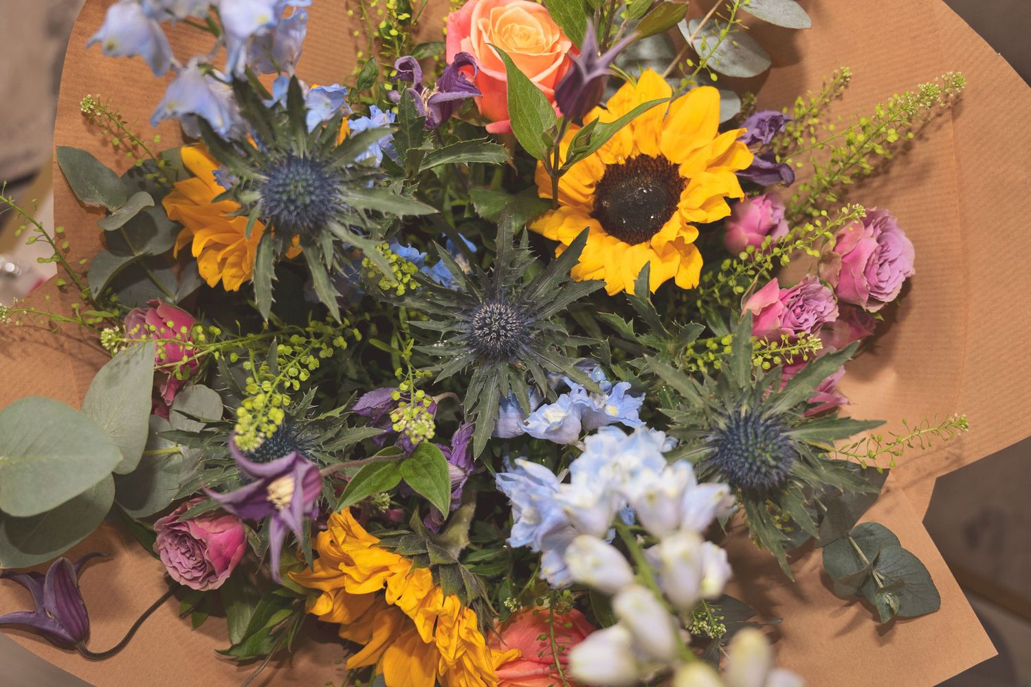 Vibrant bouquet featuring sunflowers, roses, blue thistles, and wildflowers wrapped in brown kraft paper.