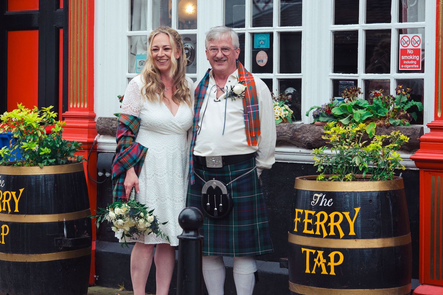 Couple posing outside the Ferry Tap pub, man wearing traditional Scottish tartan kilt, woman in white wedding dress holding bouquet.