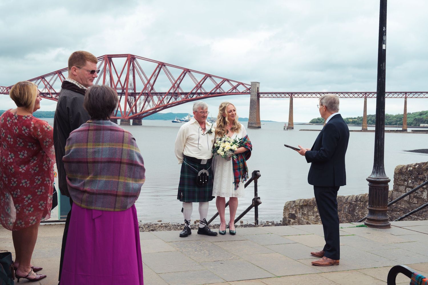 Outdoor wedding ceremony by the Forth Bridge in Scotland, with groom in kilt and guests gathered near the water.