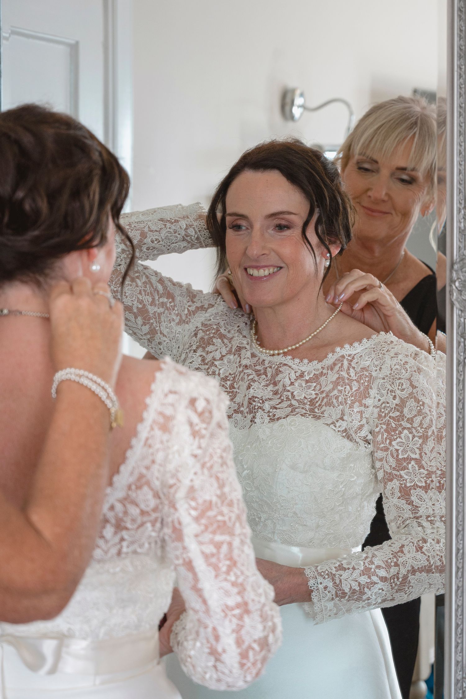 Bride in lace wedding dress getting necklace fastened by woman, smiling at mirror reflection.