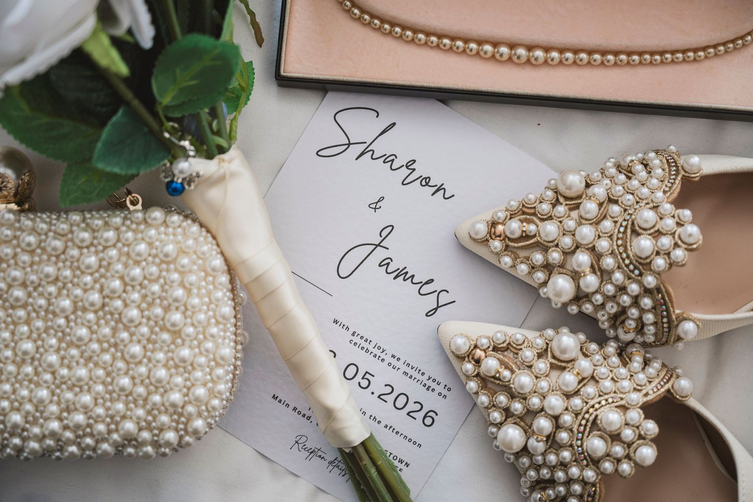 Elegant wedding flat lay with pearl-embellished heels, beaded clutch, floral bouquet, and Sharon & James 2026 invitation.