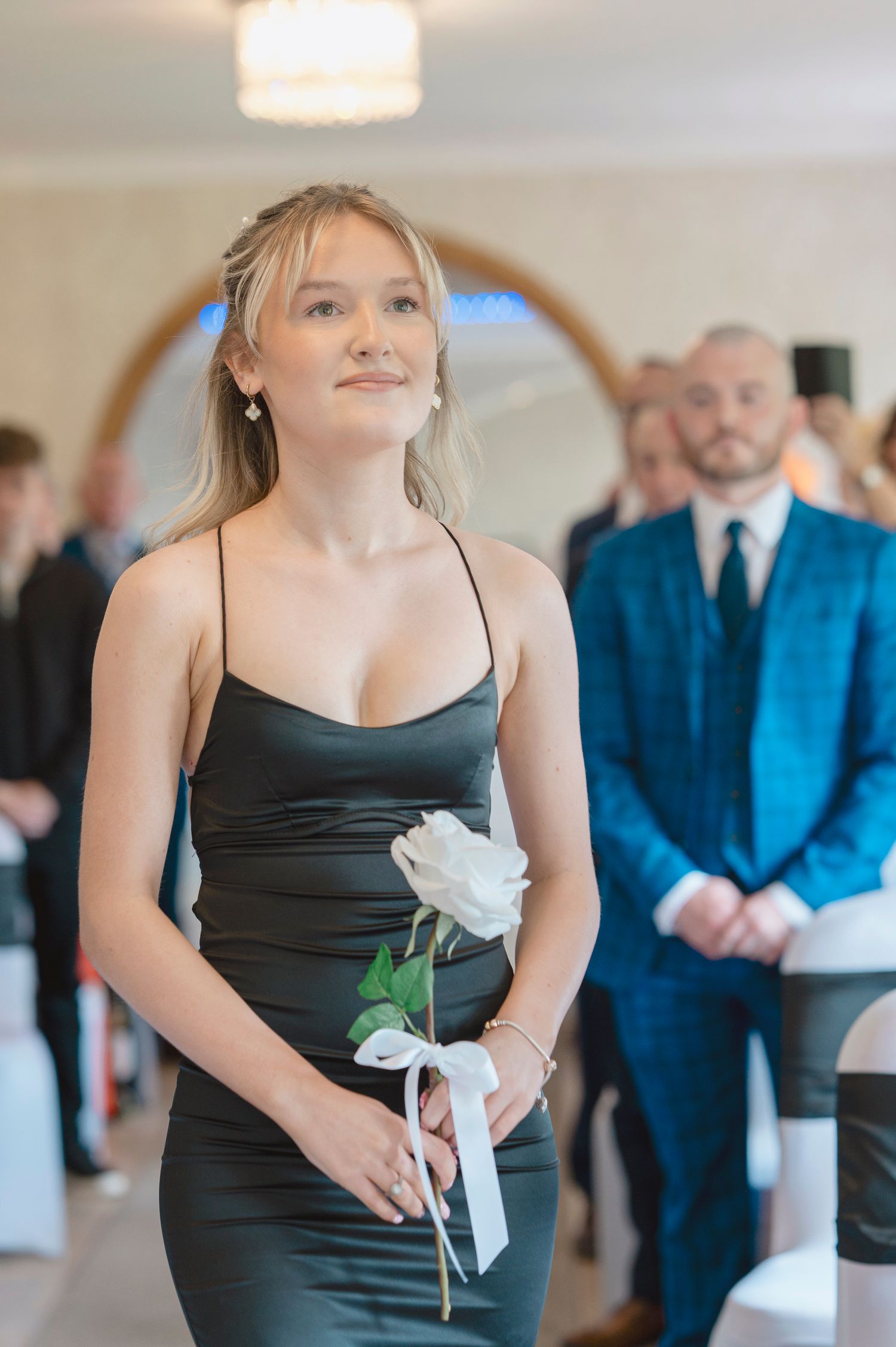 Elegant bridesmaid in black satin dress holding white rose walks down aisle at wedding ceremony.