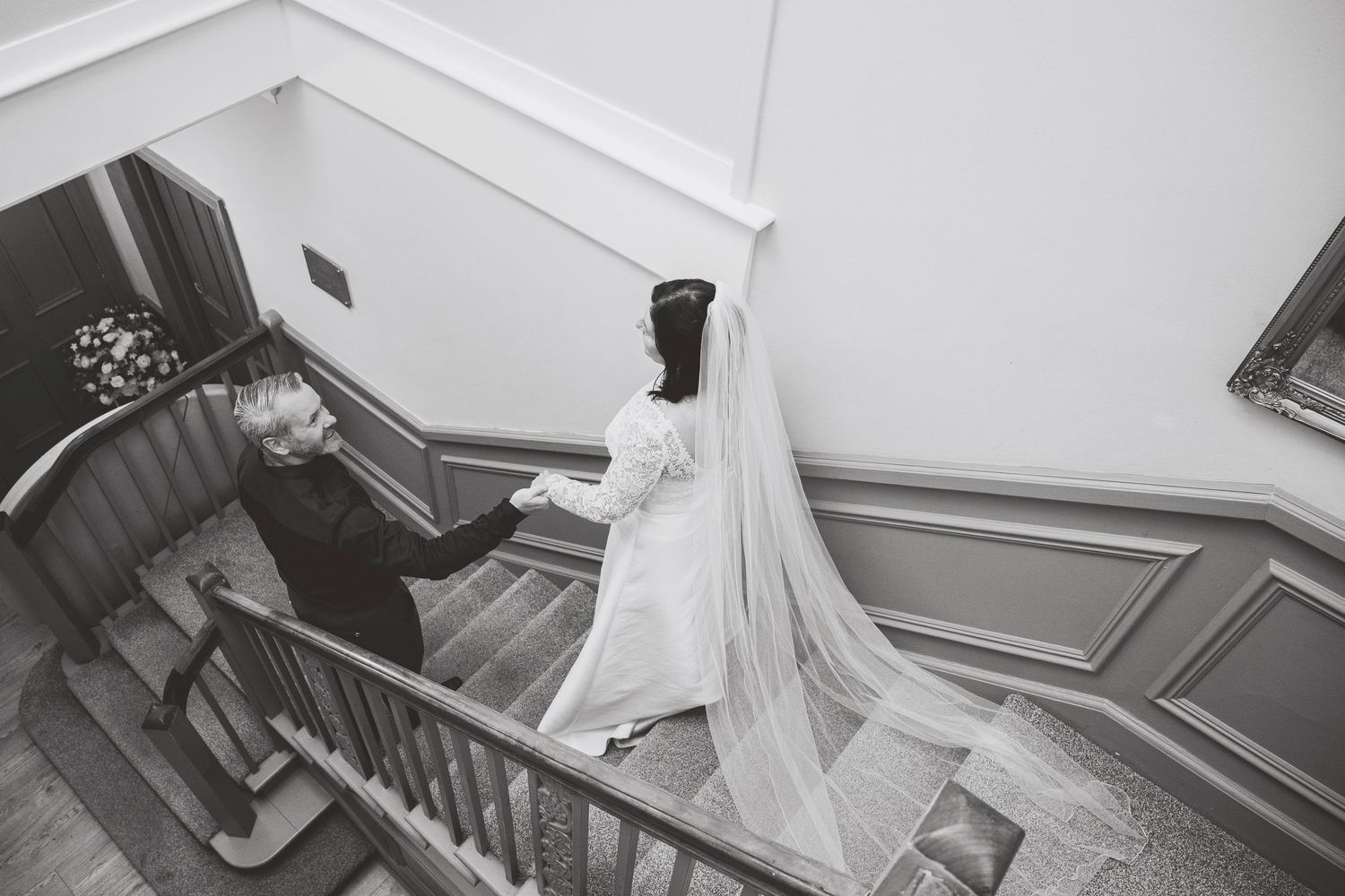 Black and white photo of a bride in a flowing veil descending an elegant staircase with a companion at a wedding venue.