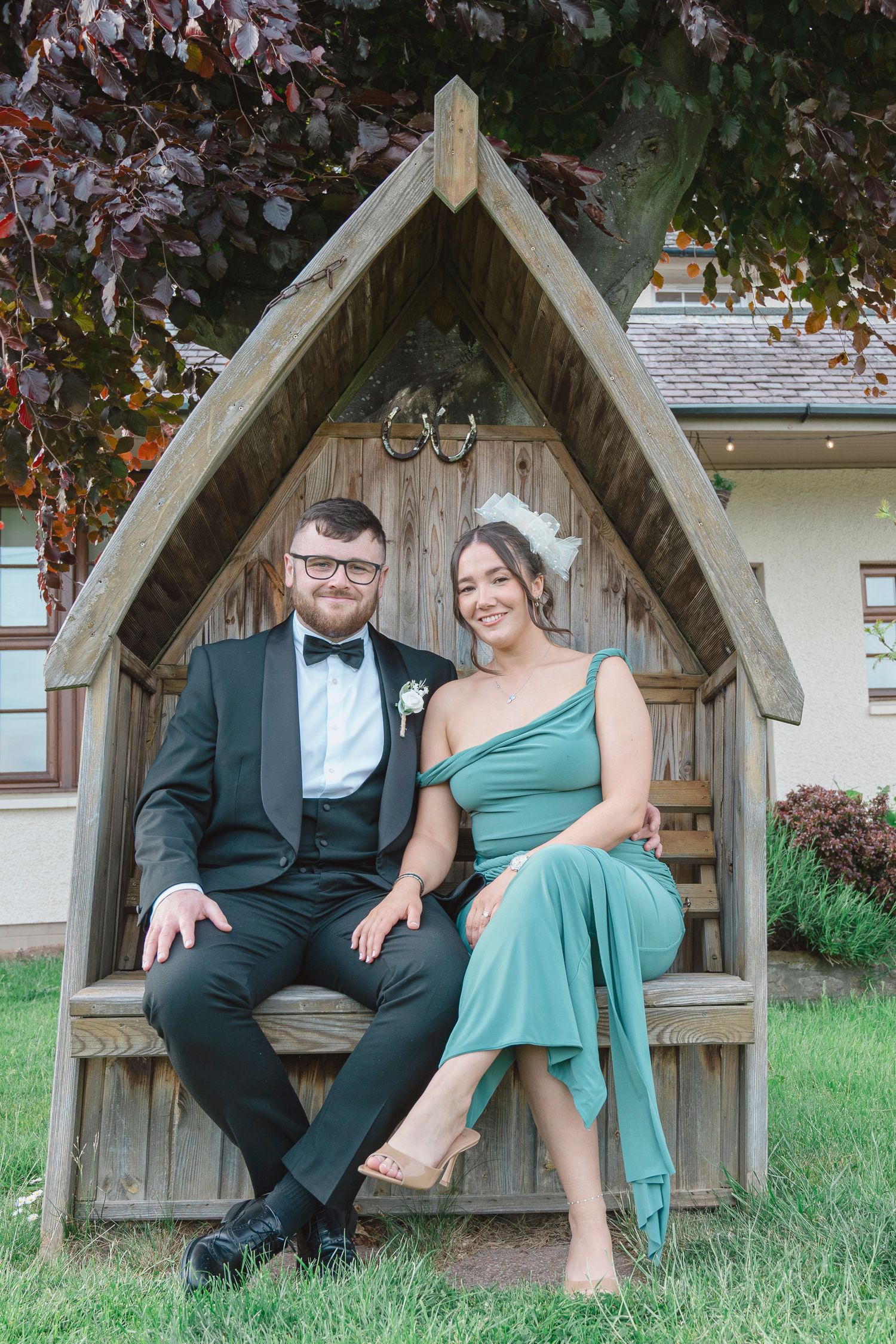 Couple in formal attire sitting in a wooden arch garden seat, man in black tuxedo, woman in green dress.