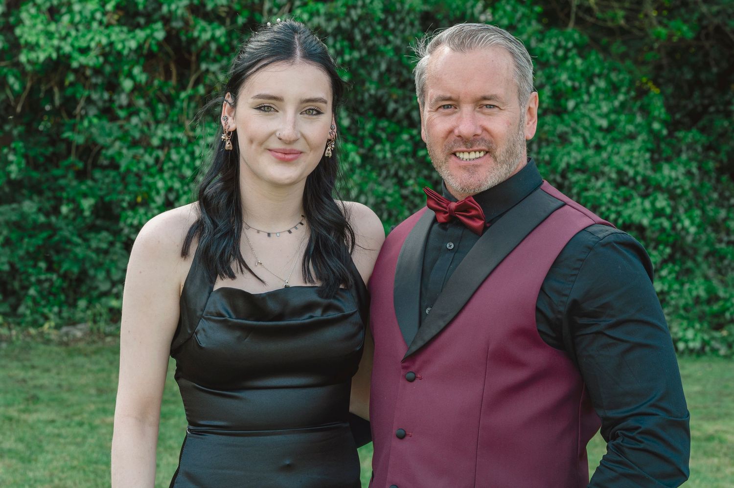 A man and woman dressed in formal attire pose together outdoors, smiling, with green foliage in the background.