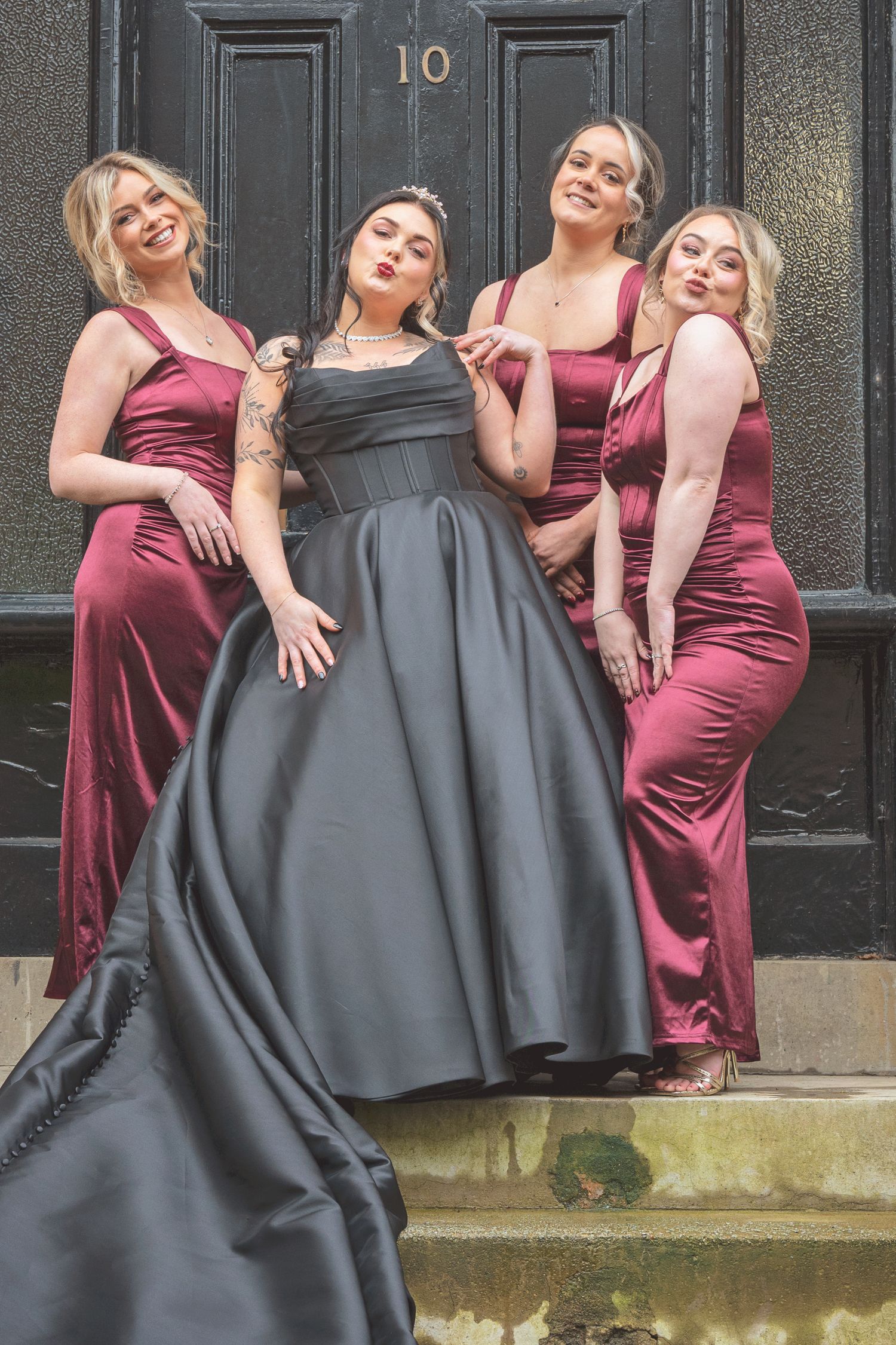 Bride in black gown strikes a fun pose with three bridesmaids in burgundy dresses outside a black door numbered 10.