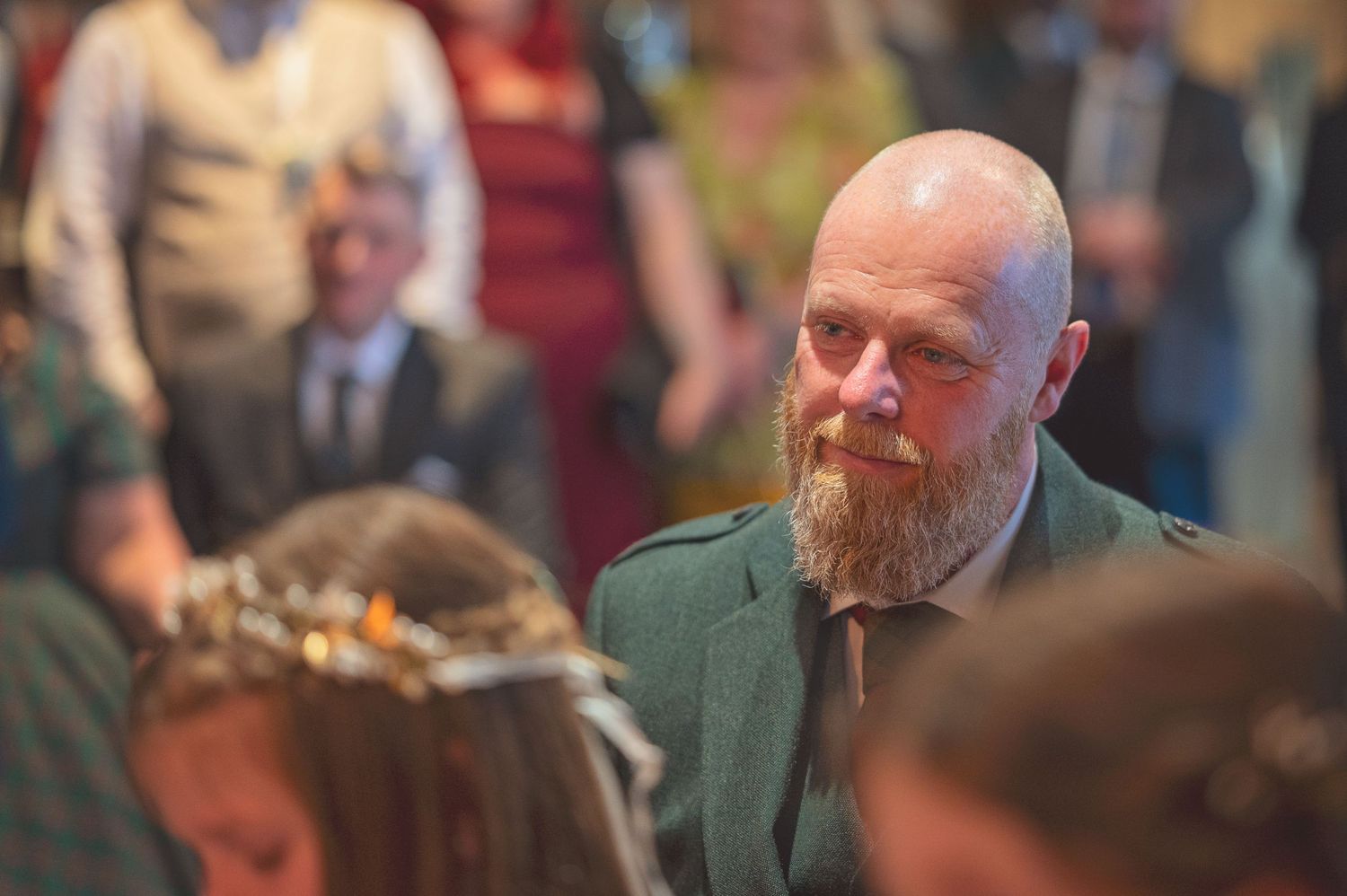 Bald bearded man in green tartan jacket watching wedding ceremony among seated guests.