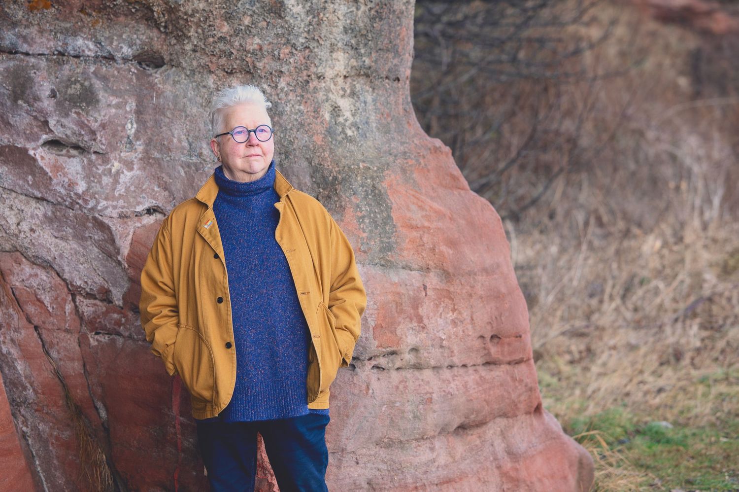 The Author Val Mcdermid with short white hair and round glasses wearing a mustard jacket and blue turtleneck sweater in the Weymss caves
