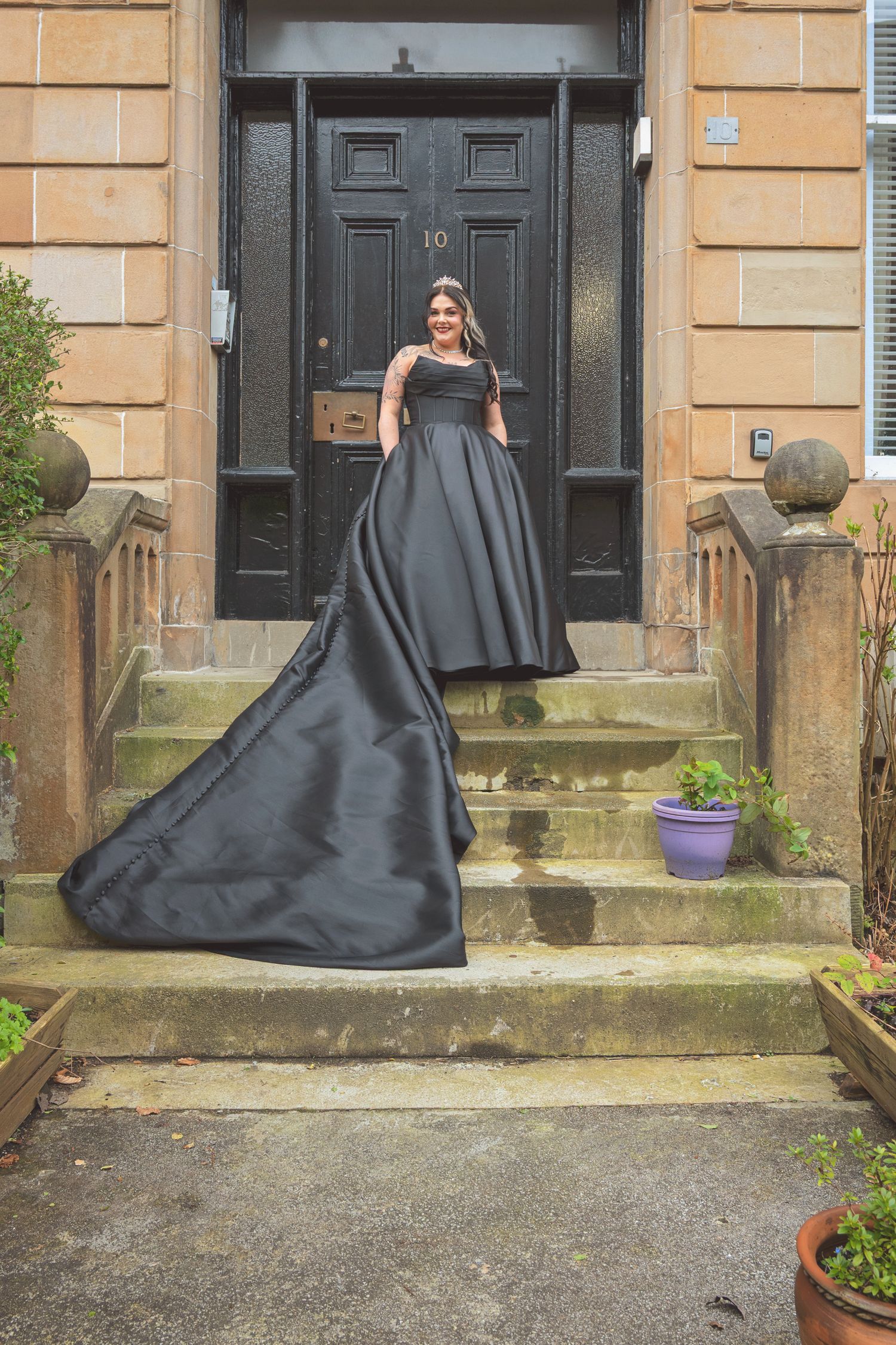Bride in dramatic black ball gown with long train poses alone on stone steps outside a black door.