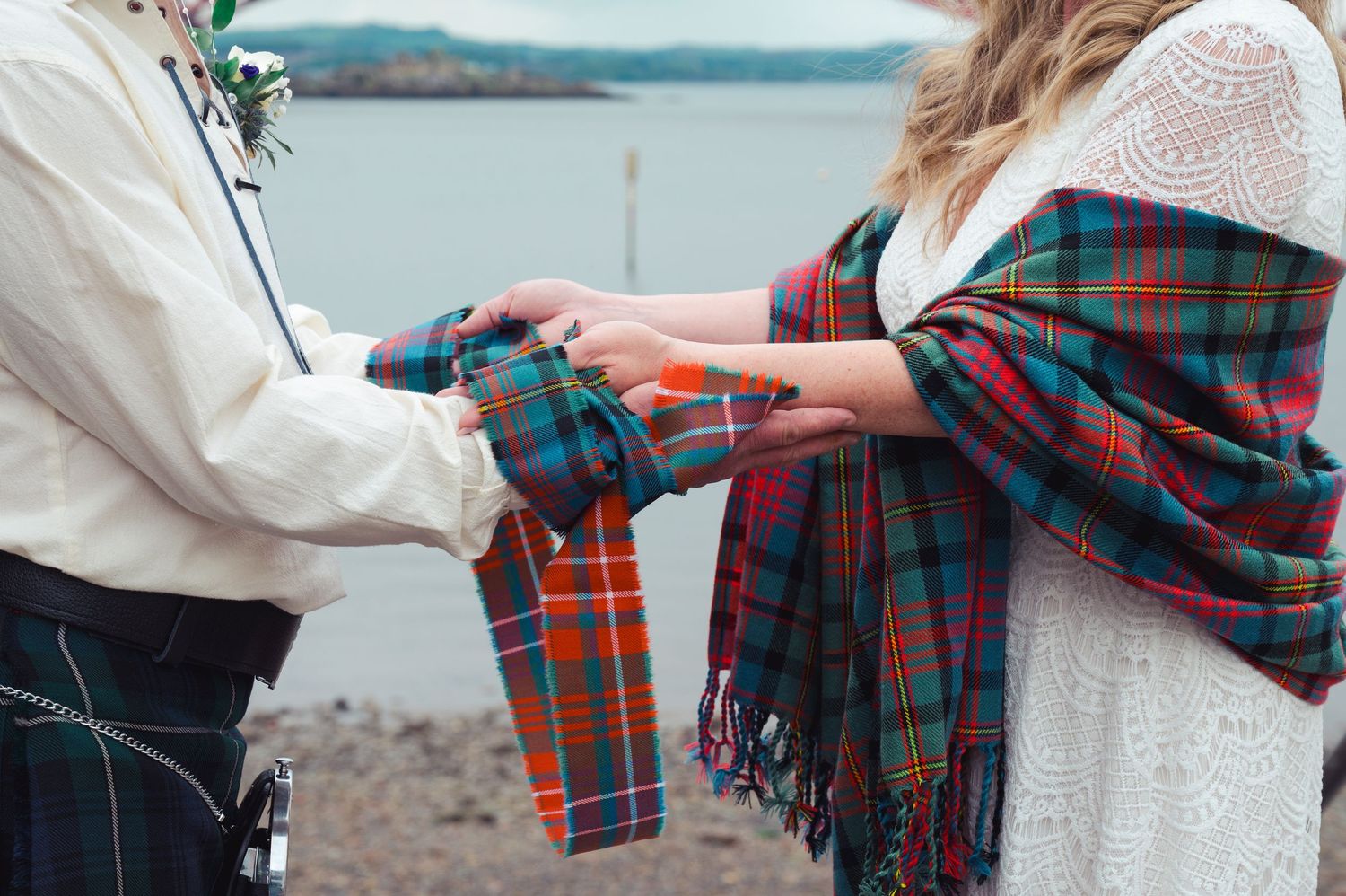 Couple performing a traditional Scottish handfasting ceremony with tartan ribbon by a loch.
