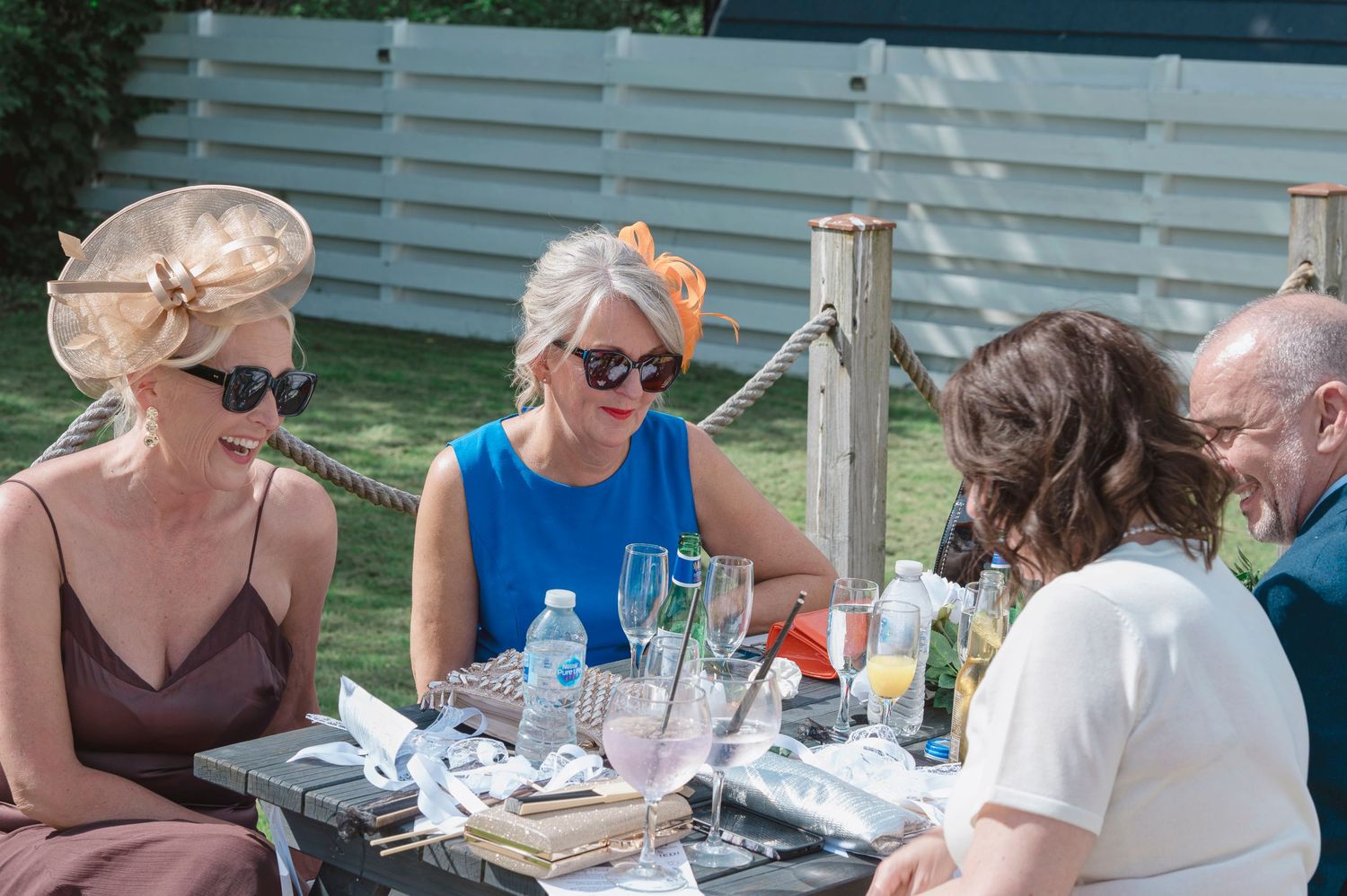 Three elegantly dressed women wearing decorative hats enjoy drinks at an outdoor table at a garden party.