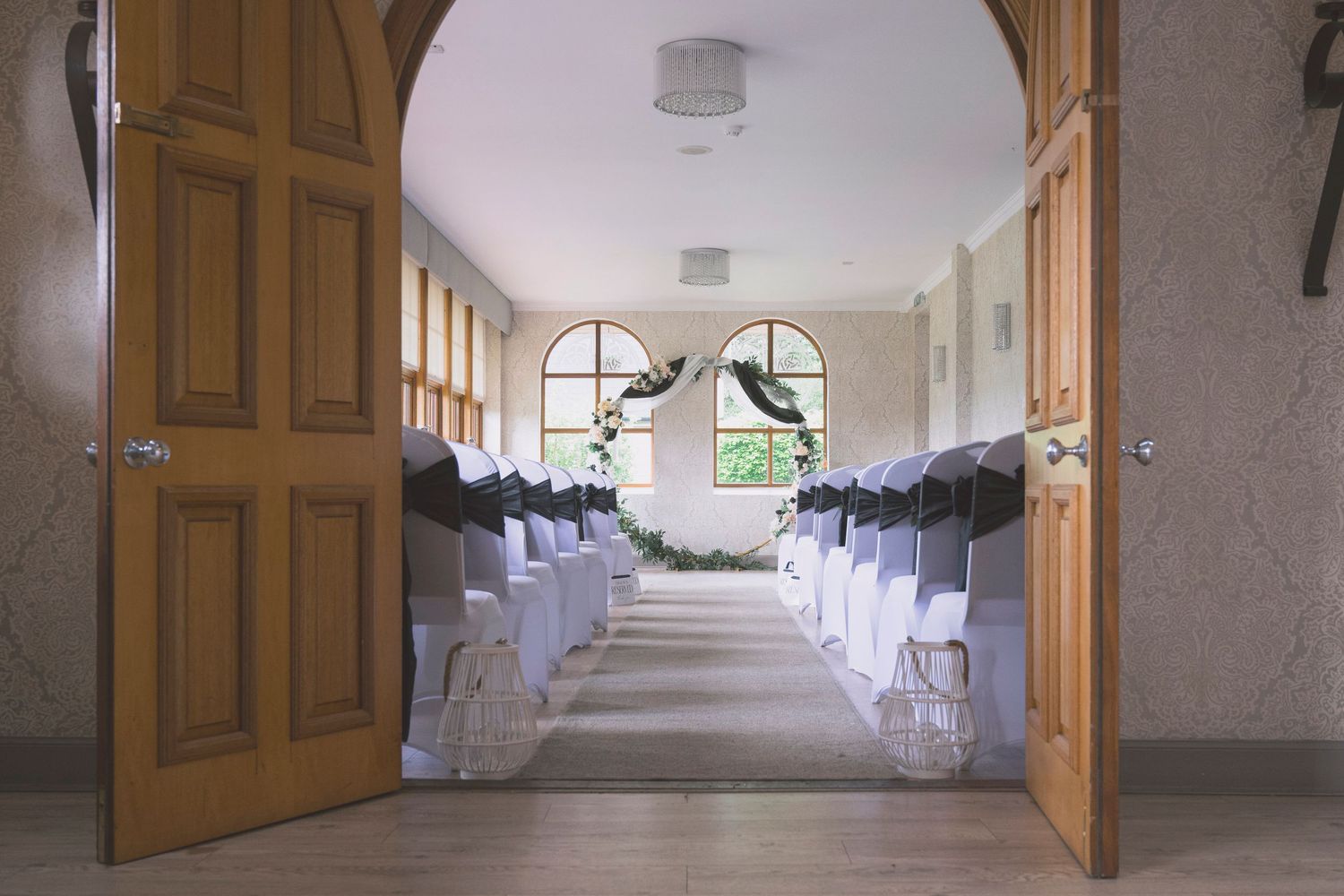 Elegant wedding ceremony venue with white chairs lining the aisle, floral decorations, and arched windows through open wooden doors.