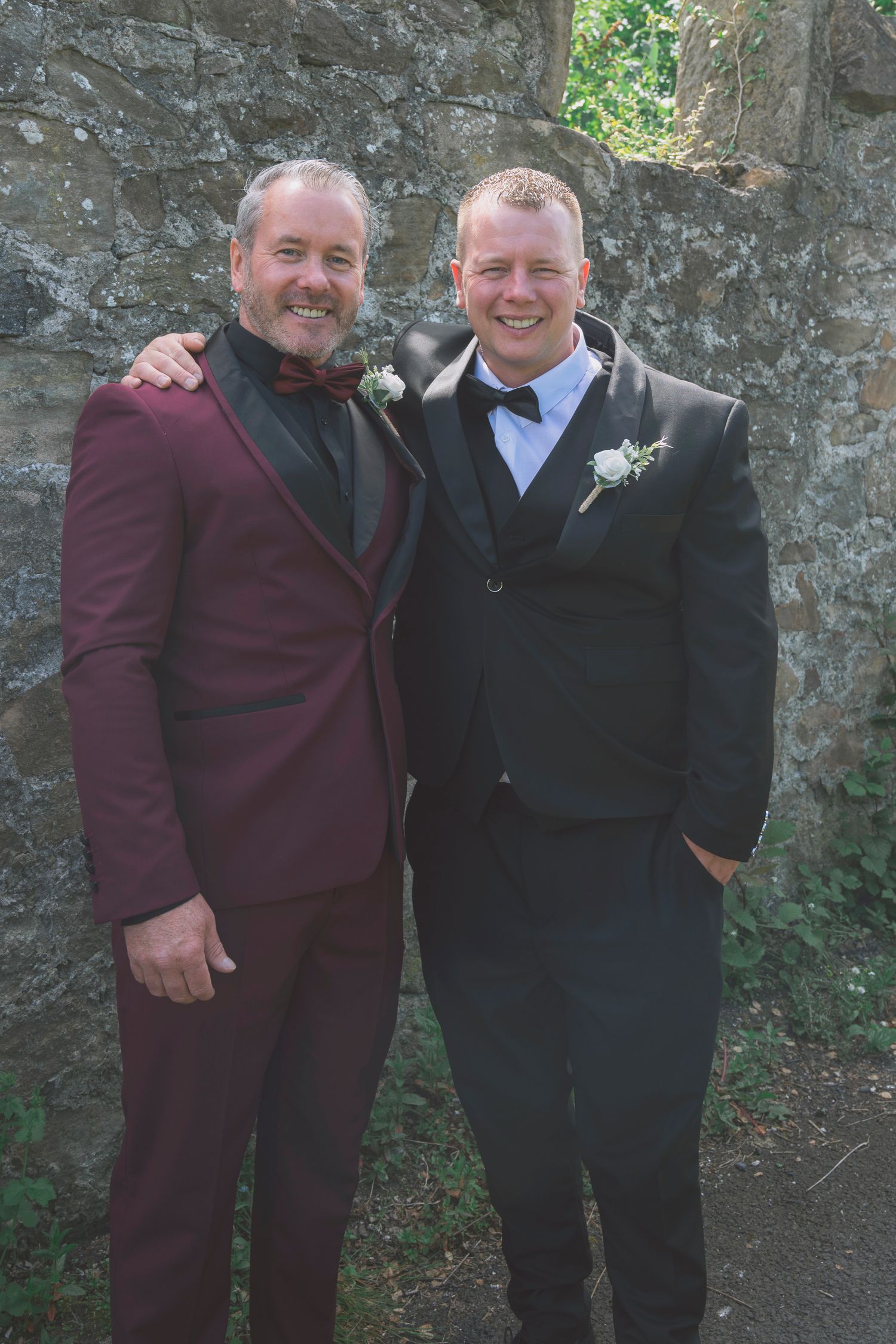 Two smiling men in formal suits with boutonnieres pose together against a stone wall at a wedding celebration.