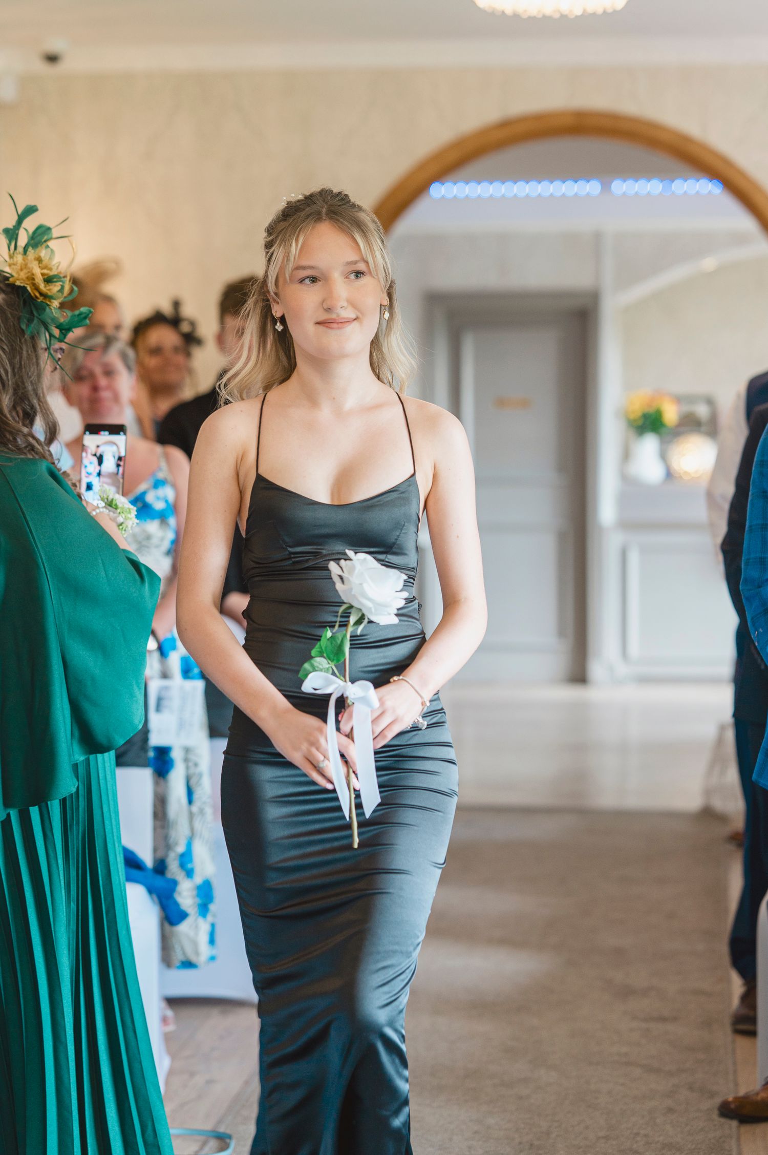 Young bridesmaid in elegant dark satin gown walks down aisle holding white rose at formal wedding ceremony.