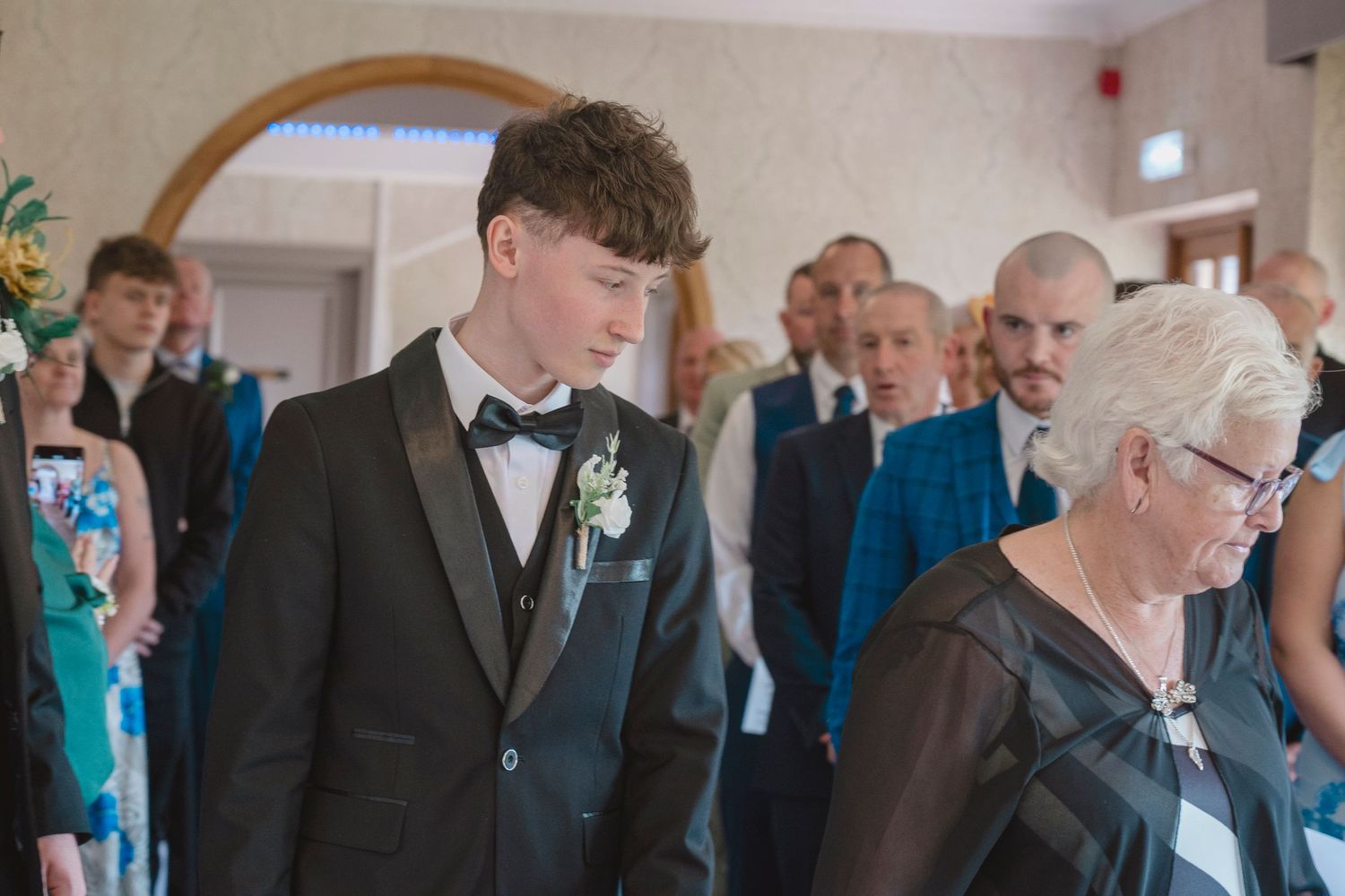 Young man in black tuxedo with boutonniere stands at a formal wedding ceremony indoors.