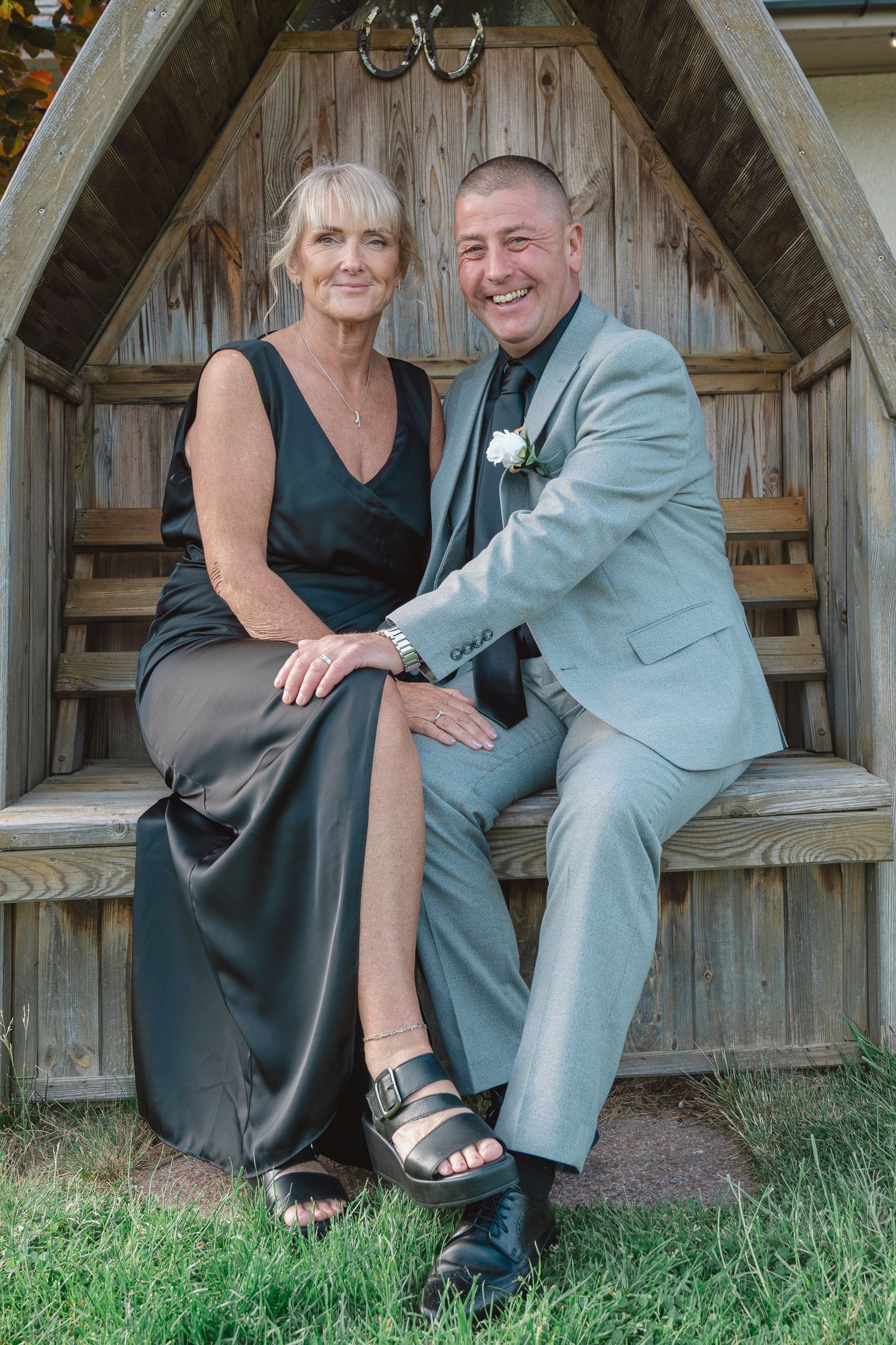 Smiling couple in formal attire sitting in a wooden gazebo, man in gray suit with boutonniere, woman in black gown.