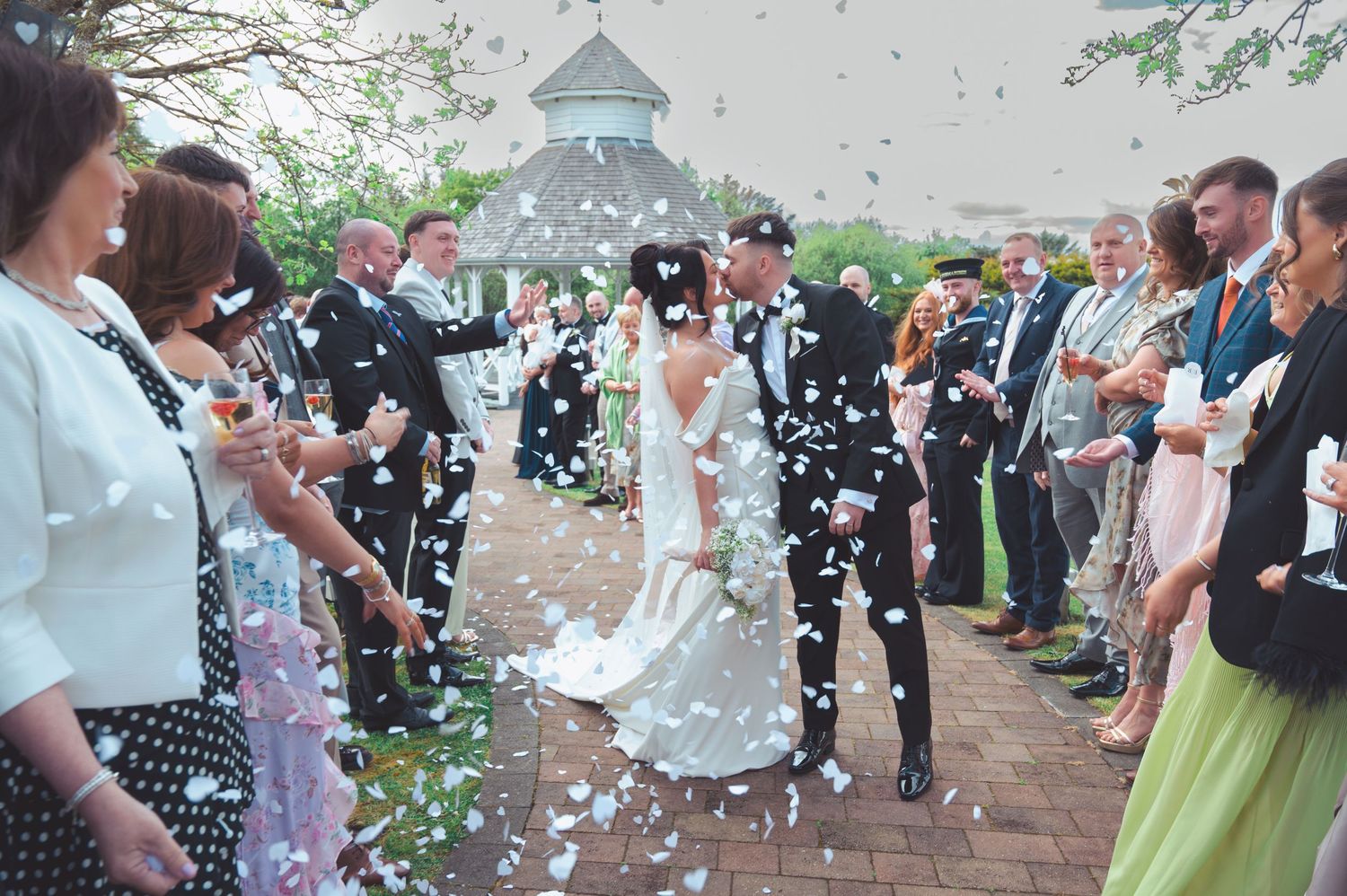 Bride and groom walk through confetti shower surrounded by cheering wedding guests outdoors.