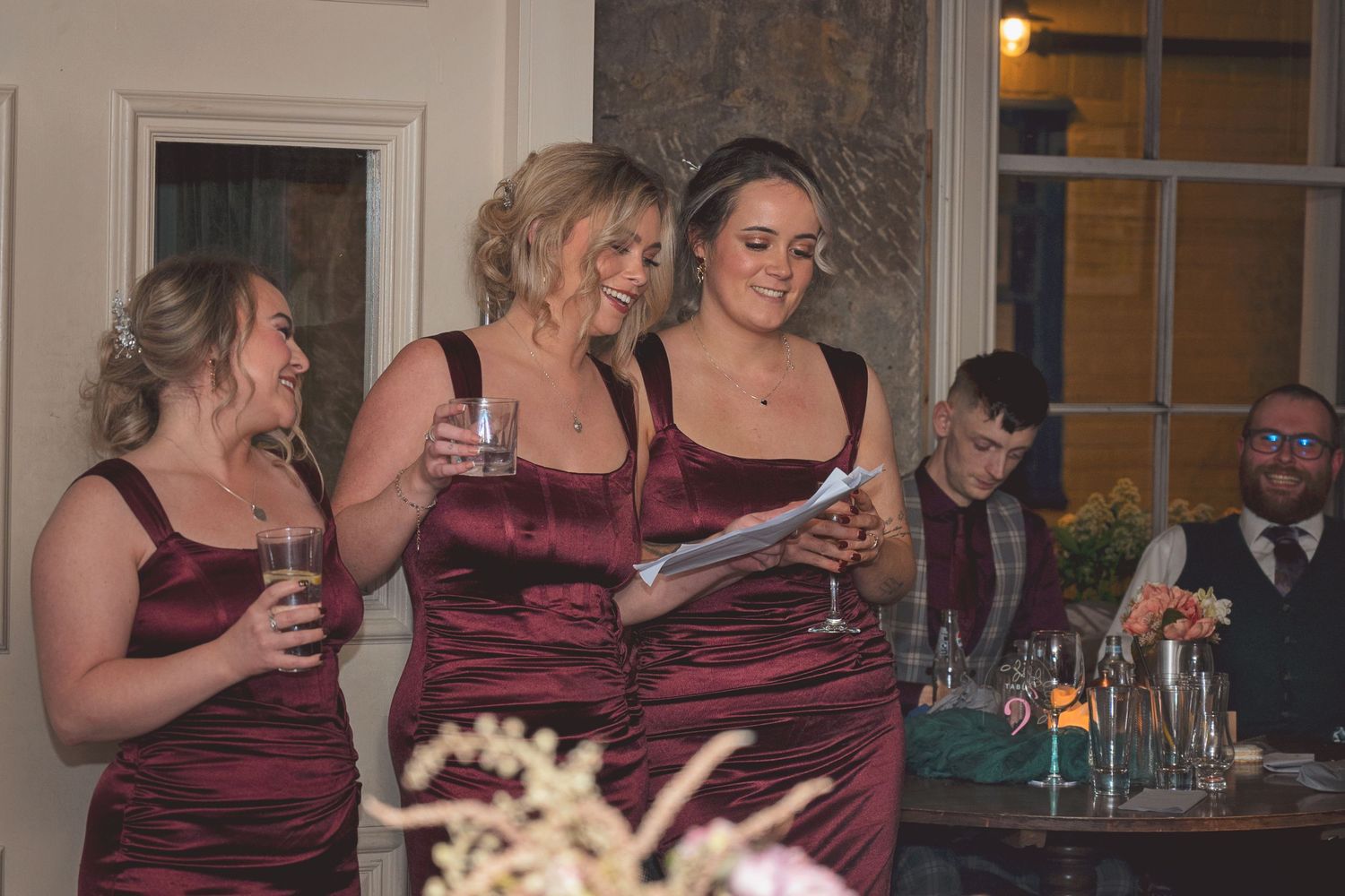 Three bridesmaids in burgundy satin dresses laugh while delivering a wedding speech.