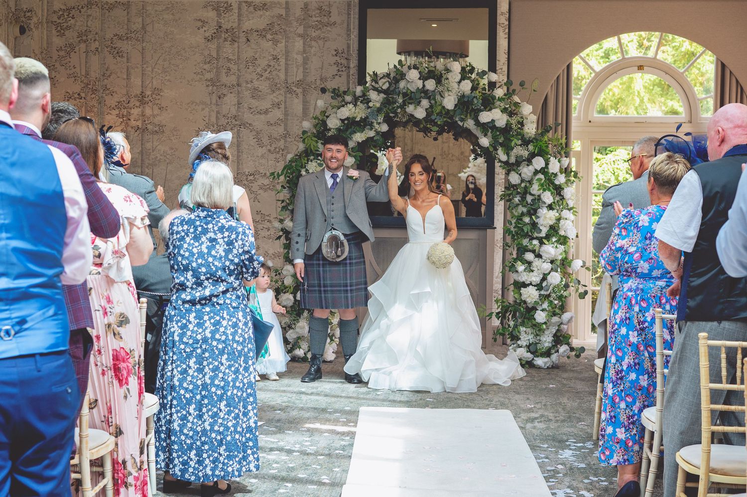 Wedding party and guests gather in a rustic venue decorated with floral arrangements.