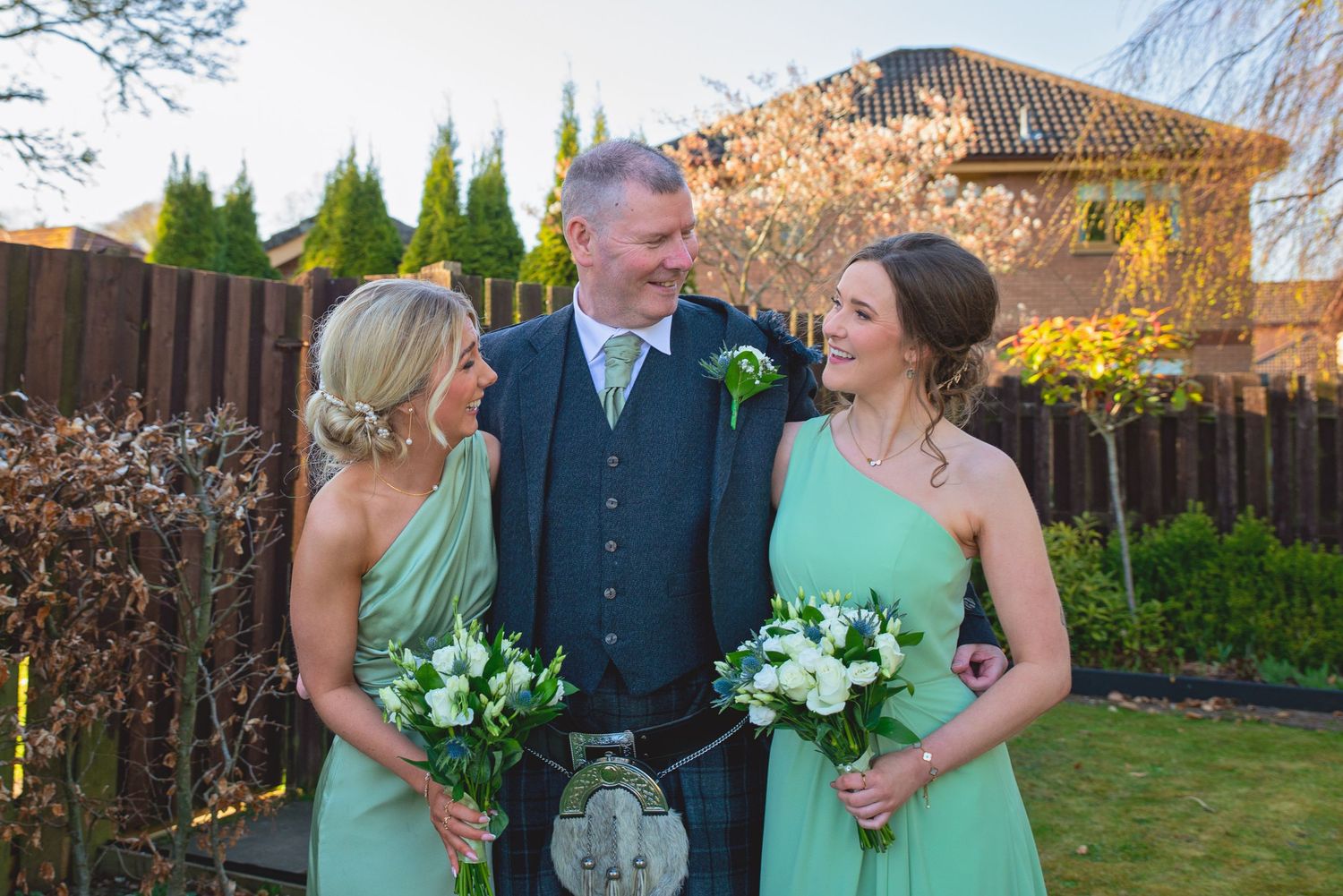 Wedding party members in mint green dresses and traditional Scottish attire pose in a garden setting.