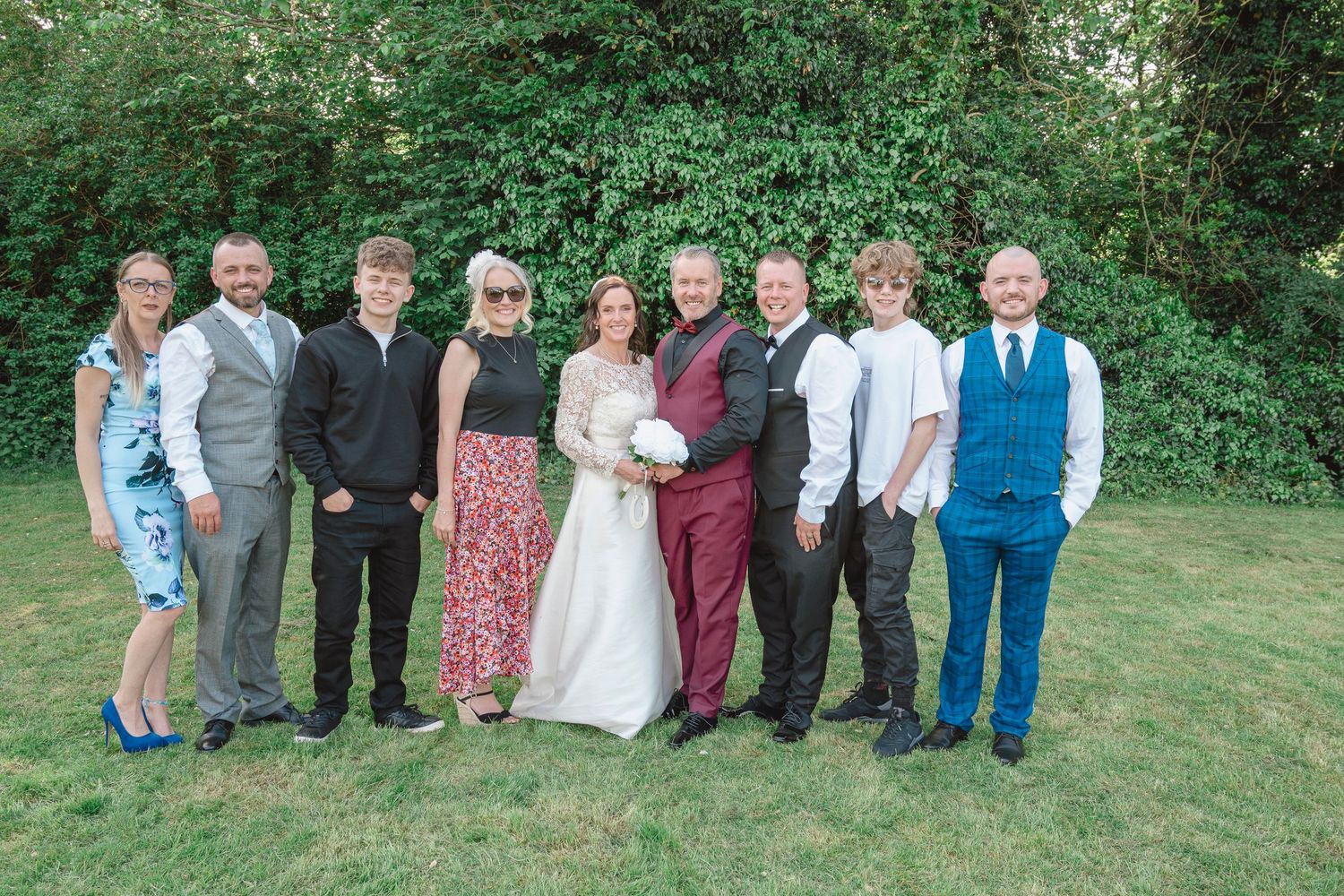 Wedding party group photo with bride and groom surrounded by family and friends on a sunny day outdoors.