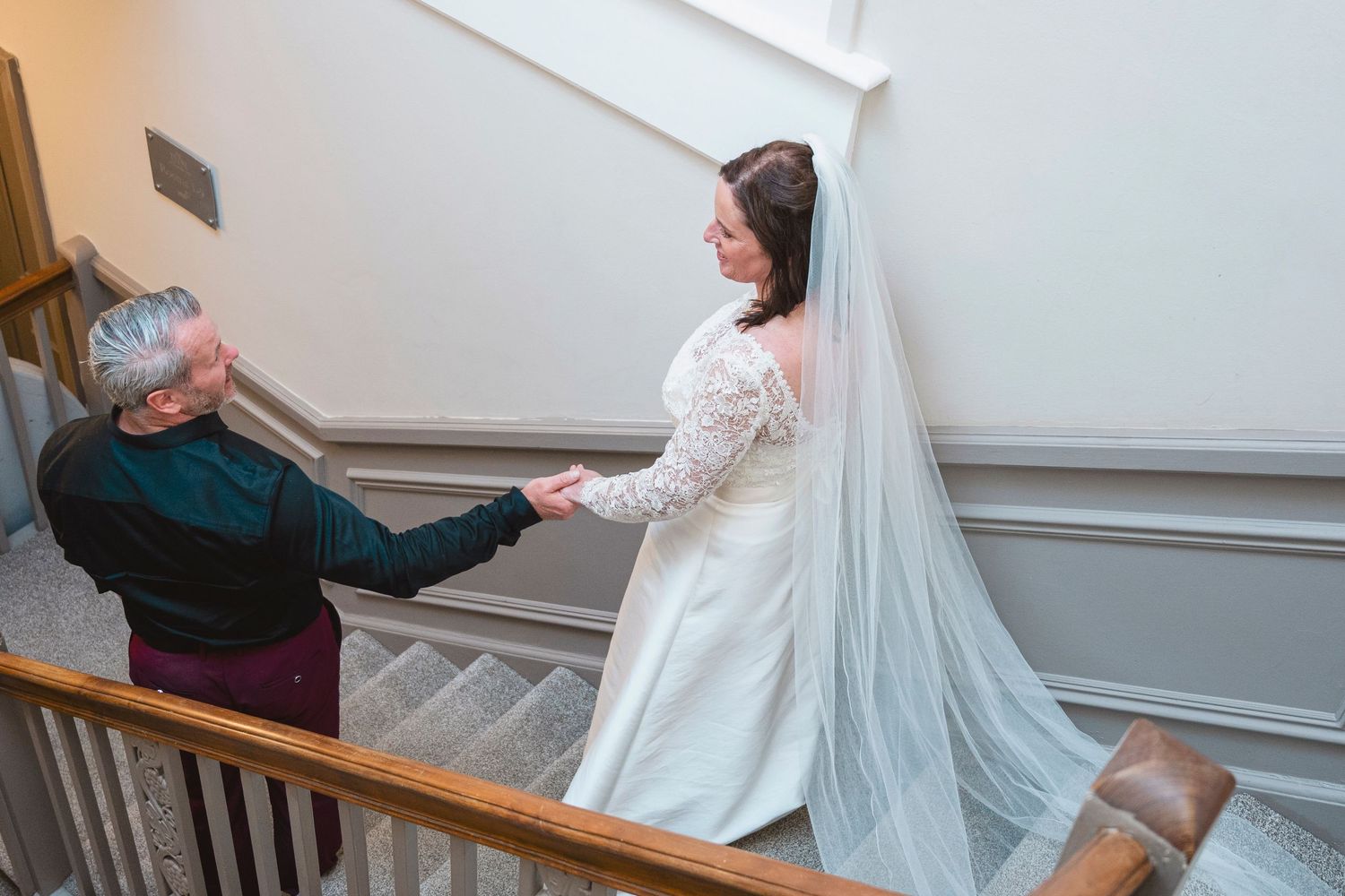 Bride in white gown and veil holds hands with guest on elegant staircase during wedding day preparations.