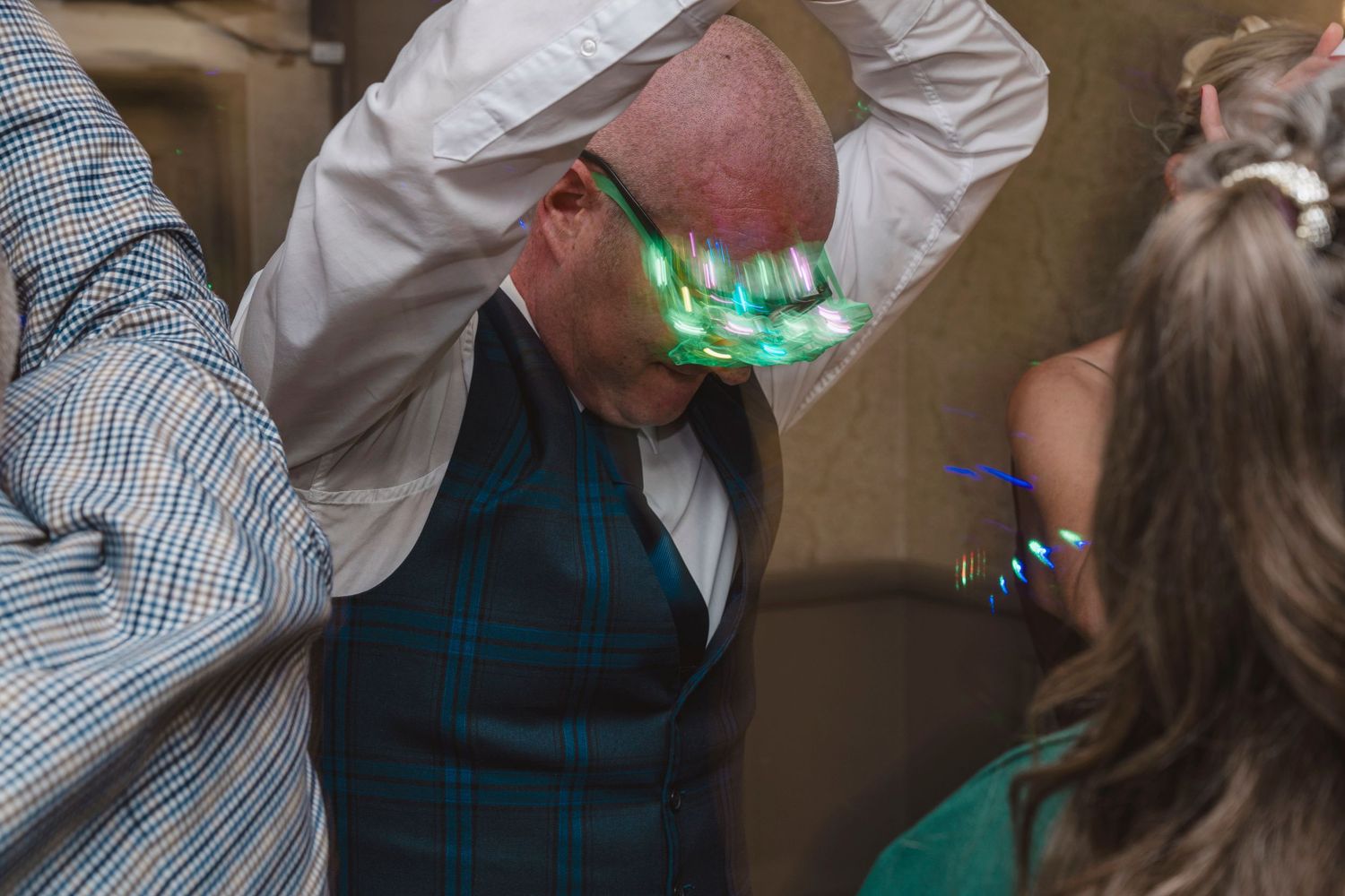 Man wearing light-up LED glasses bends forward at a lively wedding reception dance floor celebration.