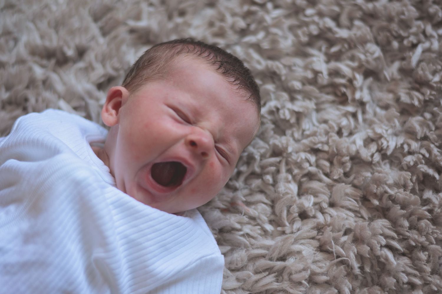 Newborn baby yawning while lying on soft beige carpet wearing white onesie.