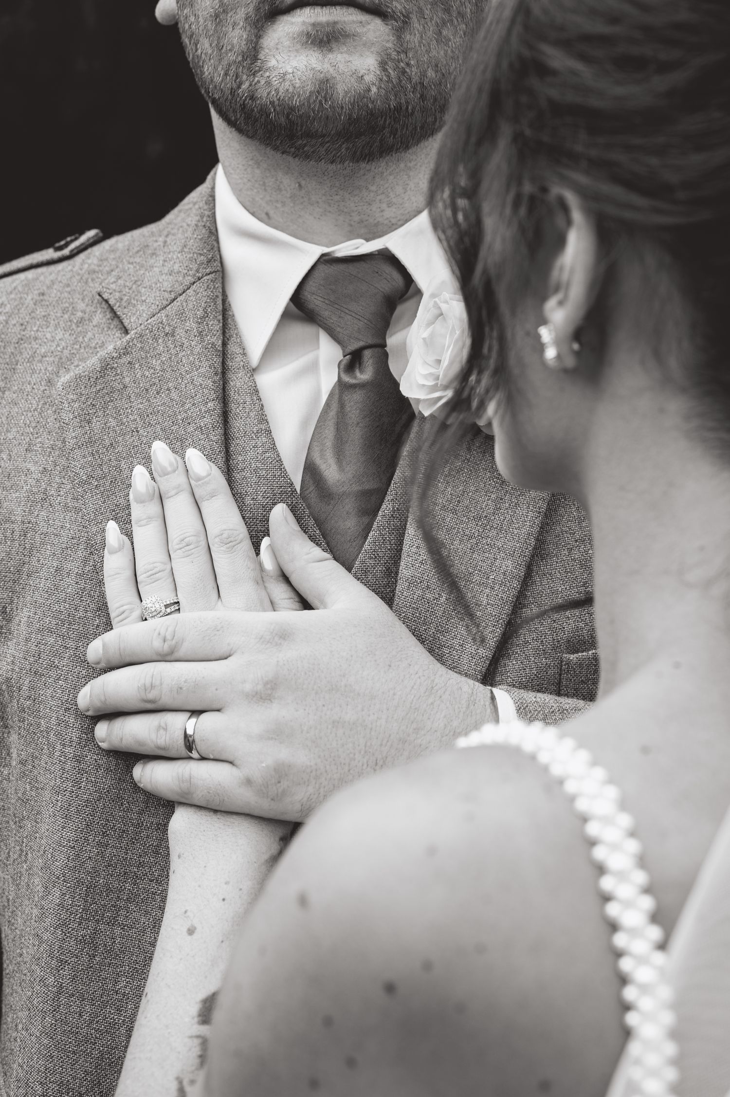 Close-up black and white detail shot of hands and wedding attire showing rings and boutonniere.