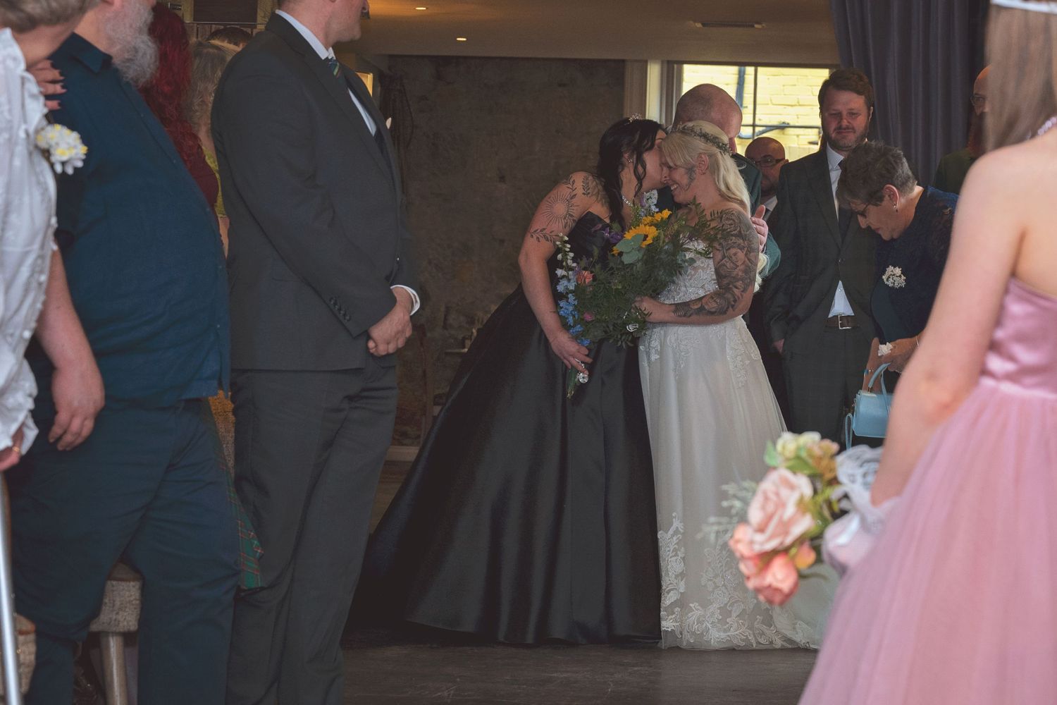 Two brides share a kiss during their wedding ceremony surrounded by guests holding bouquets.