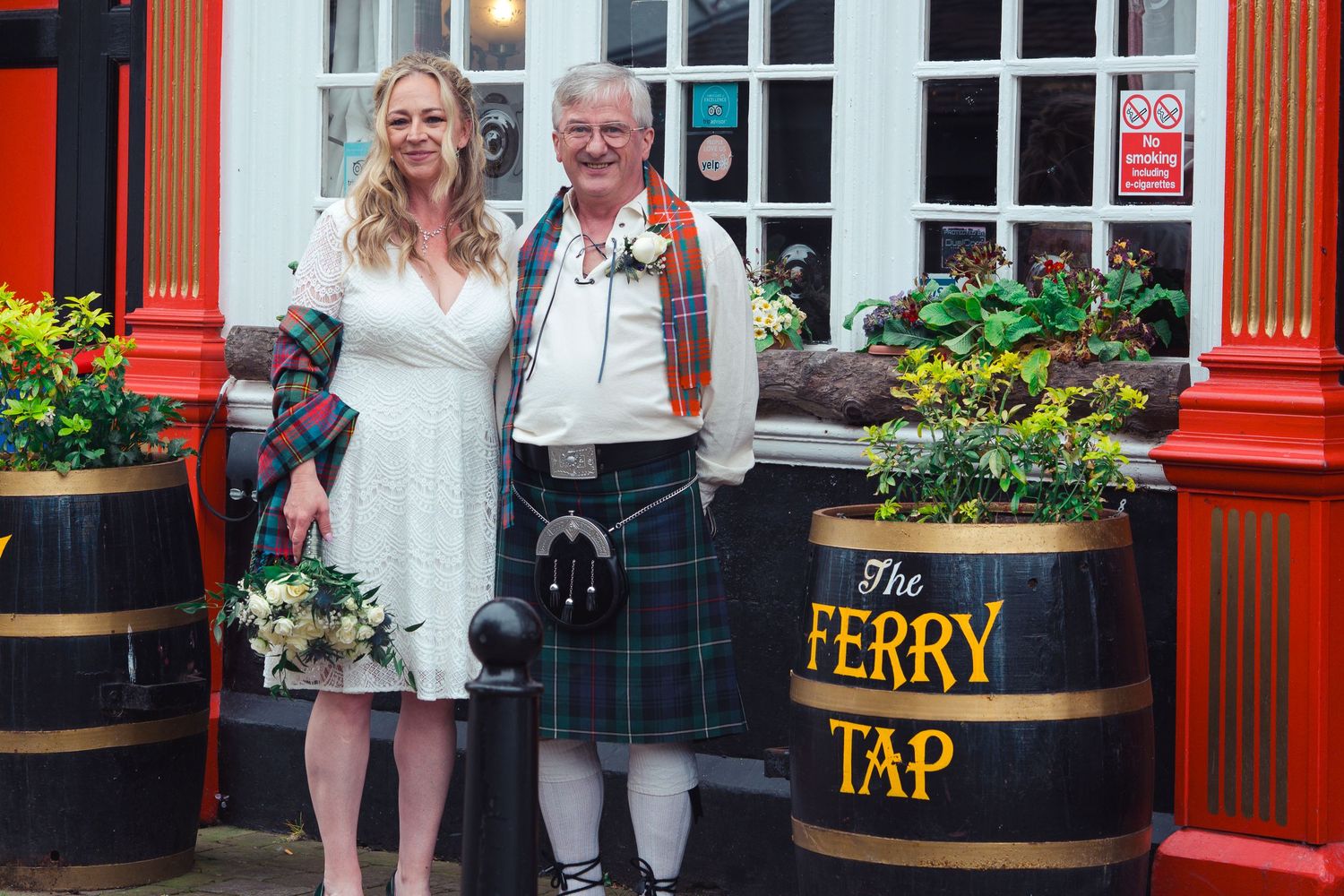 Bride and groom in Scottish kilt posing outside The Ferry Tap pub with wooden barrels and floral decorations.